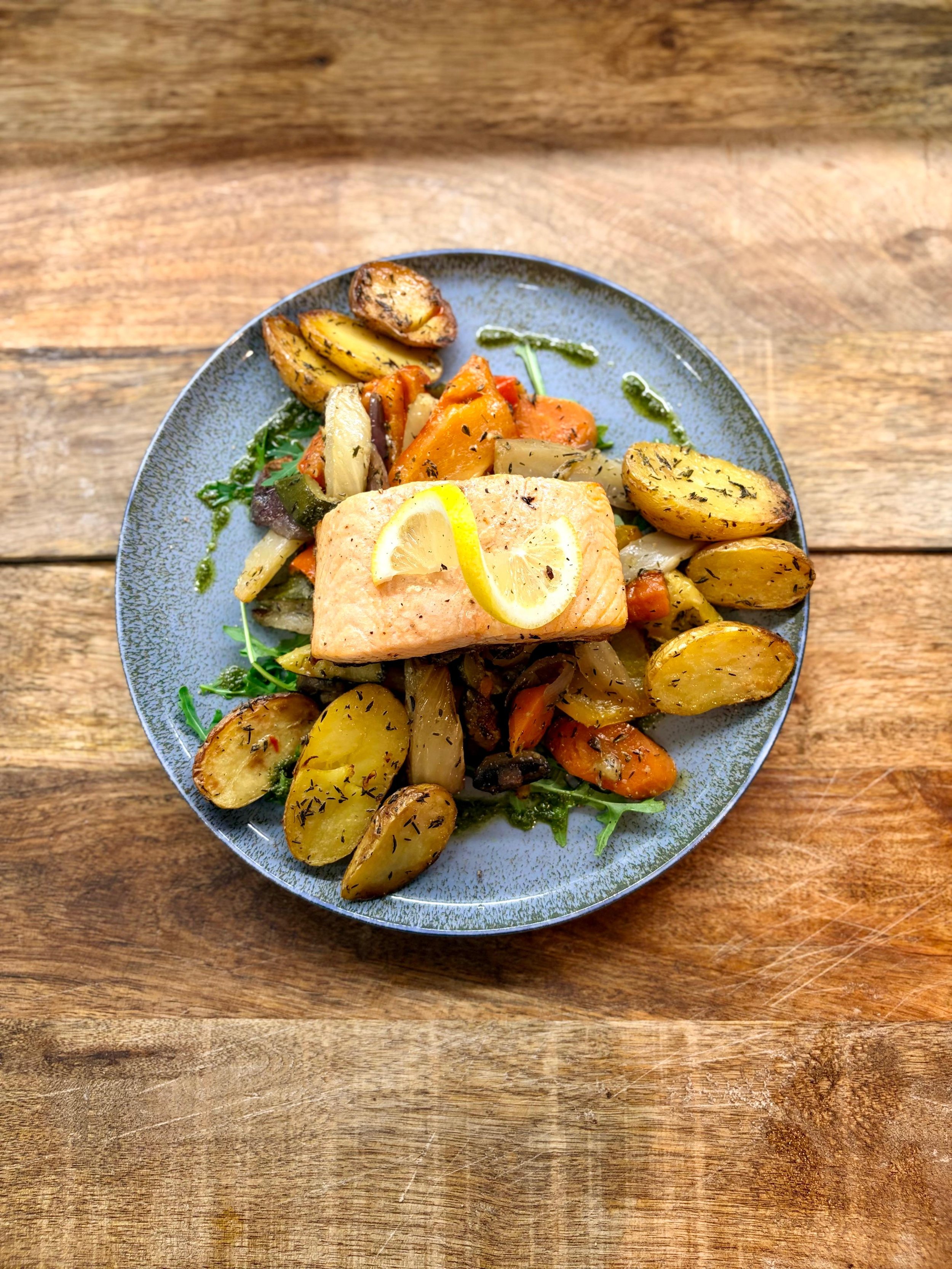 Essen im Martinus in Weingarten: Gebackener Lachs mit Zitronenscheiben auf einem Bett aus gebratenen Kartoffeln, Gemüse und Blattsalat auf einem blauen Teller auf einem Holztisch