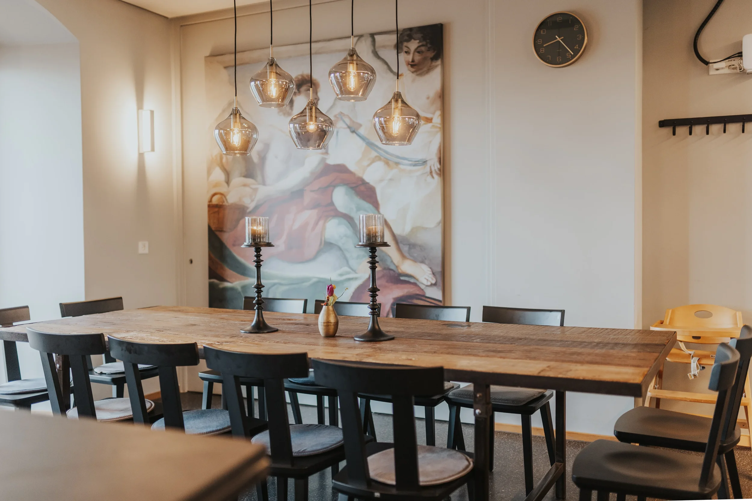 Ein stilvoll gedeckter Esstisch mit schwarzen Stühlen, Kerzenleuchtern und einer kleinen Vase, vor einer Wand mit einem großen Kunstwerk und einer Uhr, in einem modernen Raum.