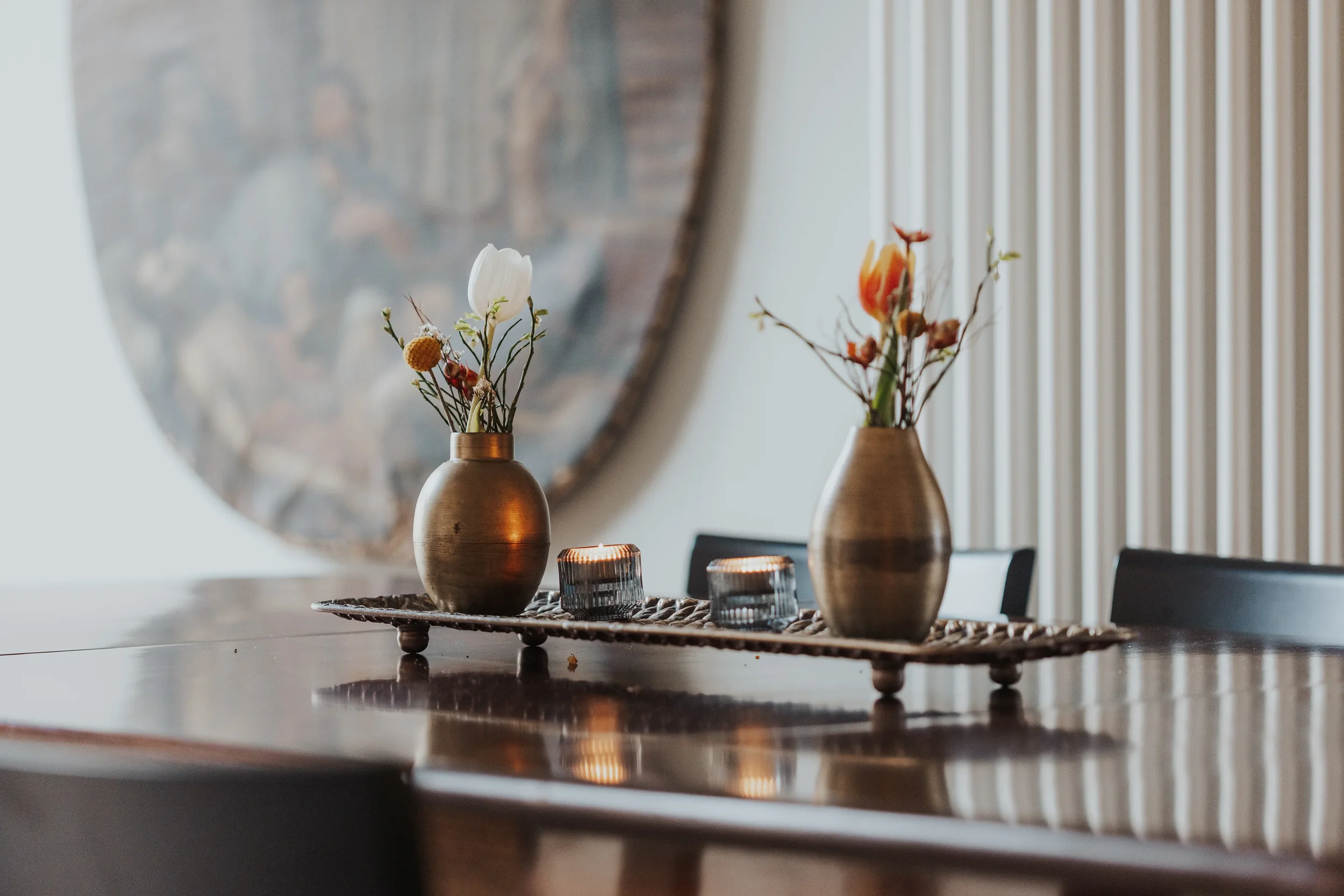Zwei bronzefarbene Vasen mit Blumen und zwei Kerzen auf einem Holztablett auf einem Esstisch, im Hintergrund vertikale Holzpaneele