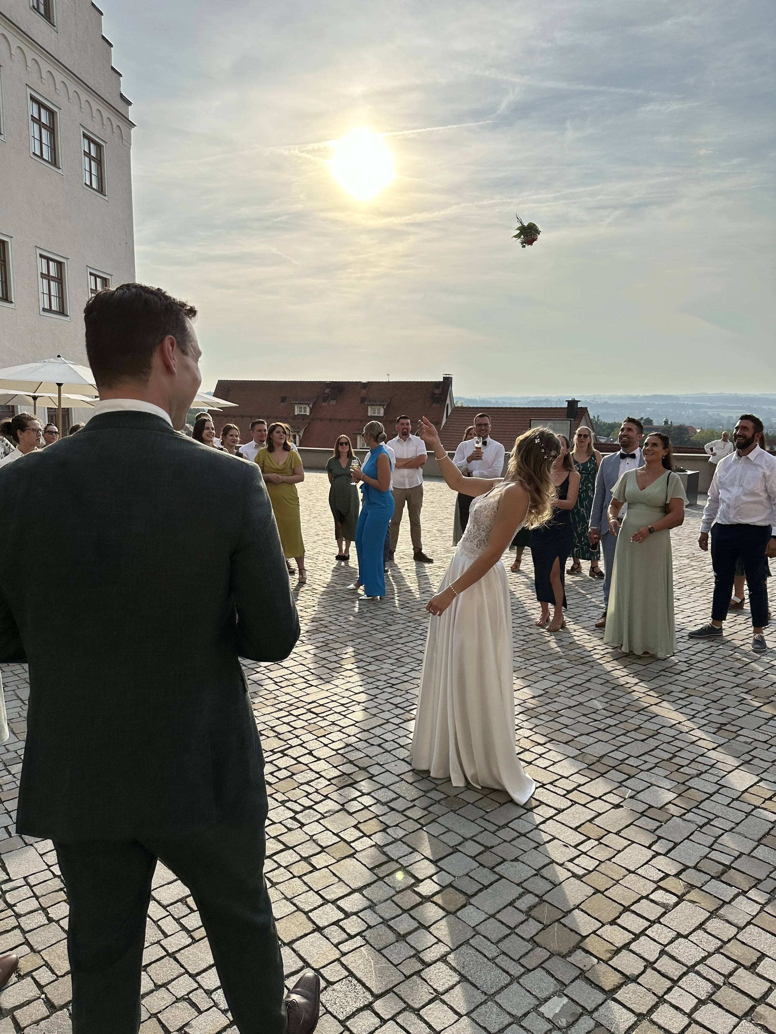 Hochzeitsparty im Freien bei Sonnenuntergang vor dem Martinus in Weingarten, mit Braut in weißem Kleid, die den Brautstrauß wirft, vor vielen Gästen auf einer Steinterrasse.
