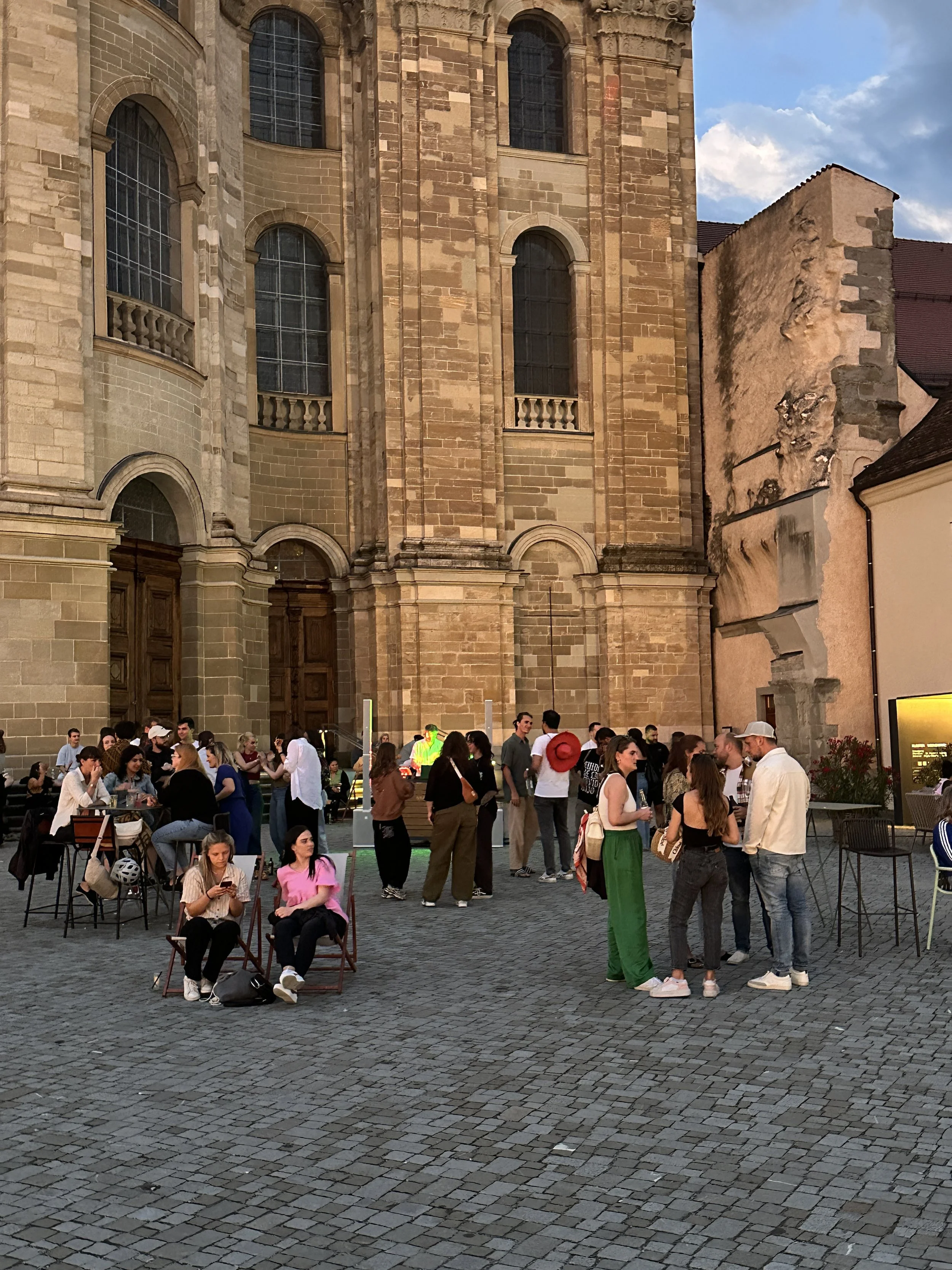 Menschen versammelt sich vor dem Martinus bei der Basilika St. Martin bei Sonnenuntergang, einige sitzen auf Stühlen, andere stehen und sprechen.