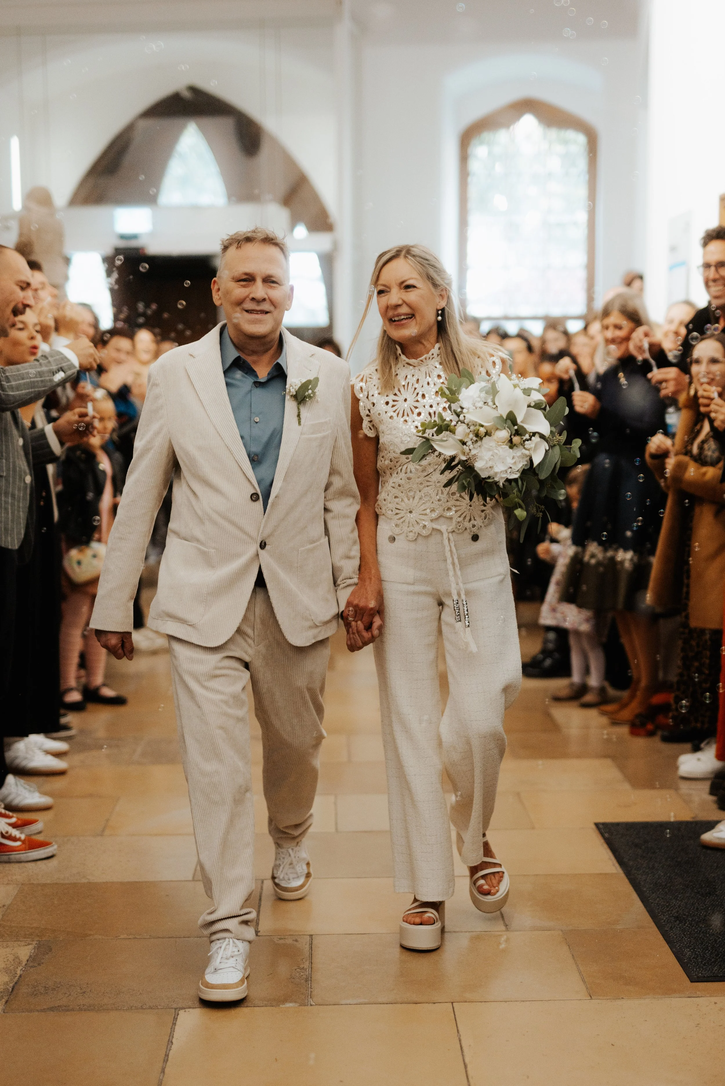 Ein Brautpaar, das Hand in Hand läuft, umgeben von jubelnden Gästen bei einer Hochzeit in einem hellen Raum.