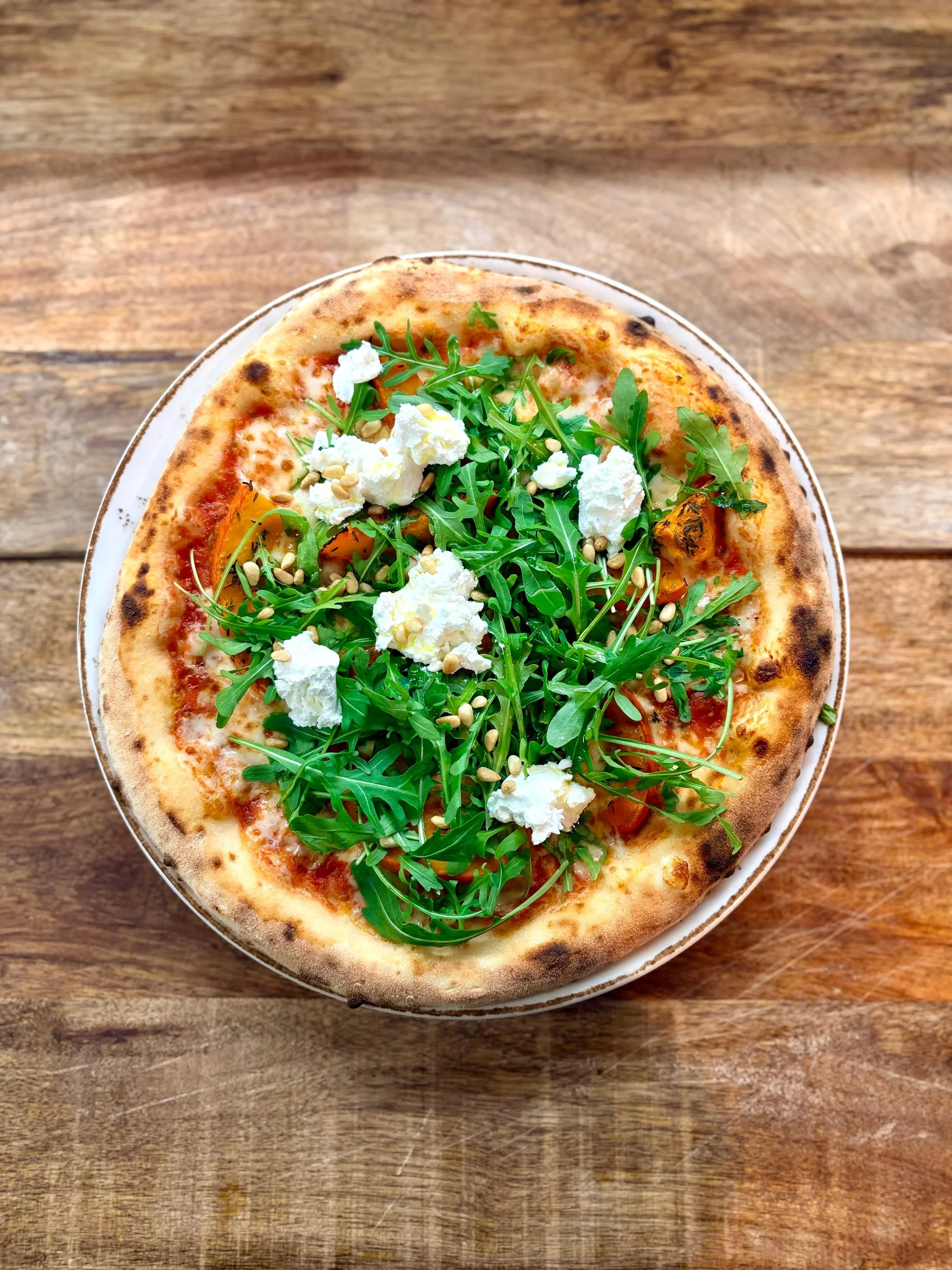 Essen im Martinus:  Pizza mit Rucola, Ricotta, Tomaten und Pinienkernen auf einem Holztisch