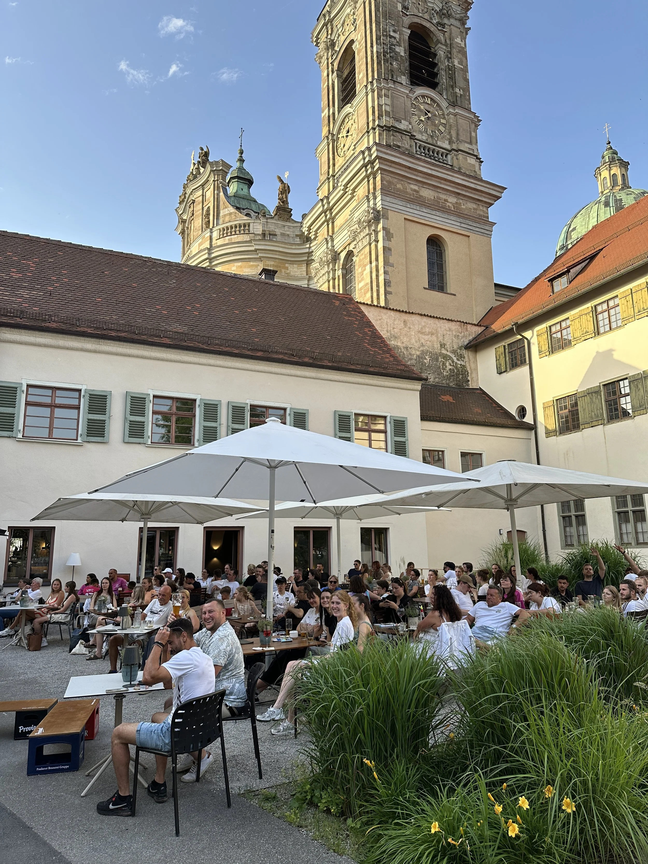 Menschen sitzen bei einem Sommerabend im Freien an Tischen unter Sonnenschirmen vor dem Martinus in Weingarten.