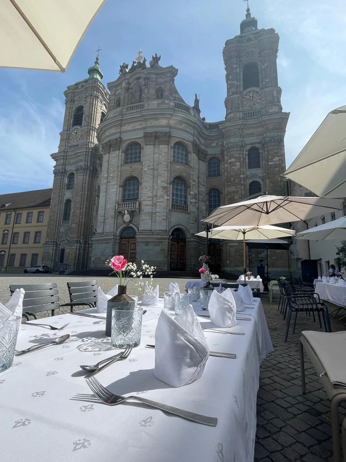 Ein gedeckter Tisch im Freien mit weißen Tischdecken, Gabeln, Messern, Gläsern und kleinen Vasen mit Blumen im Martinus in Weingarten vor der Basilika St. Martin.