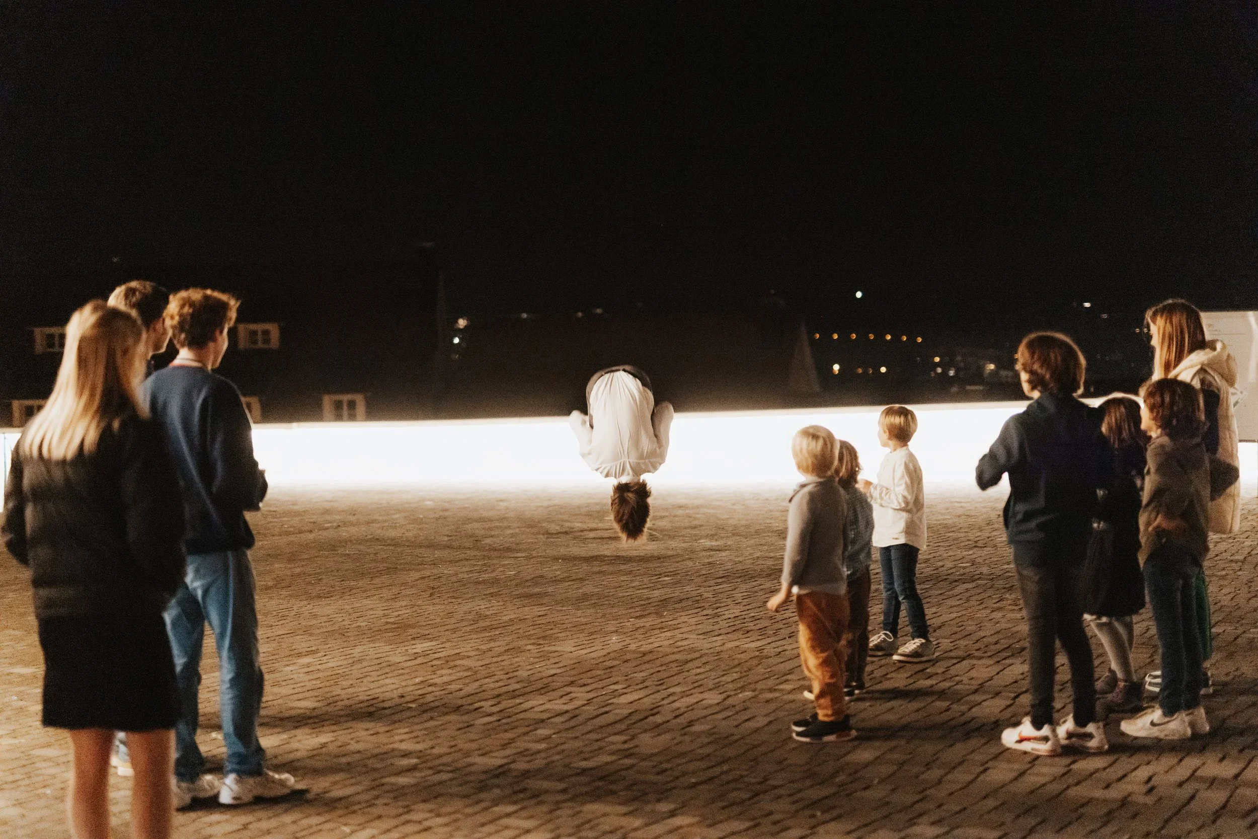 Ein Mädchen hängt kopfüber an einer leuchtenden Bar, während eine Gruppe von Menschen auf einem gepflasterten Platz bei Nacht zuschaut.