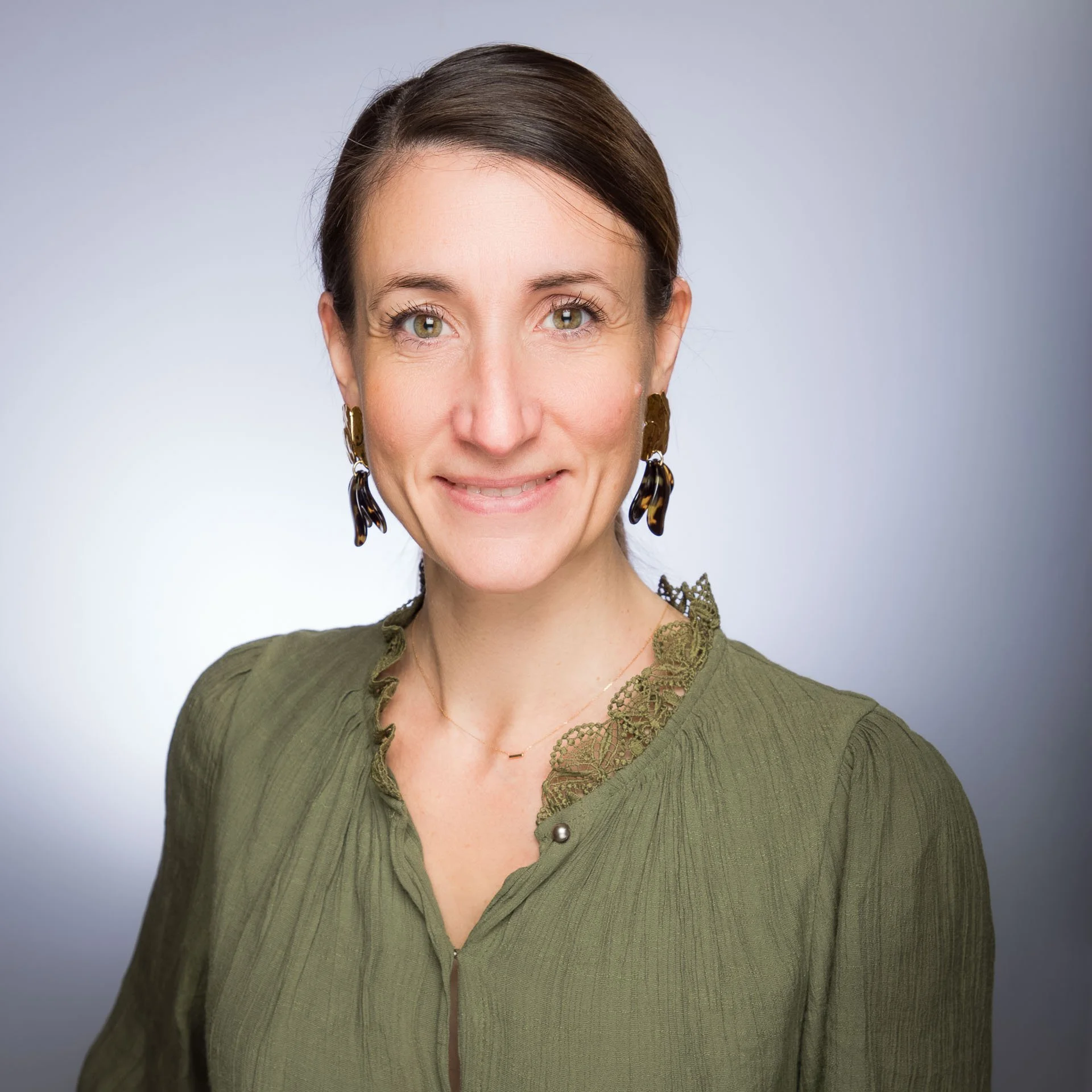 Portrait d'une femme souriante portant une blouse verte avec des détails en dentelle au col, des boucles d'oreilles en tortue et une chaîne fine, contre un fond gris clair.