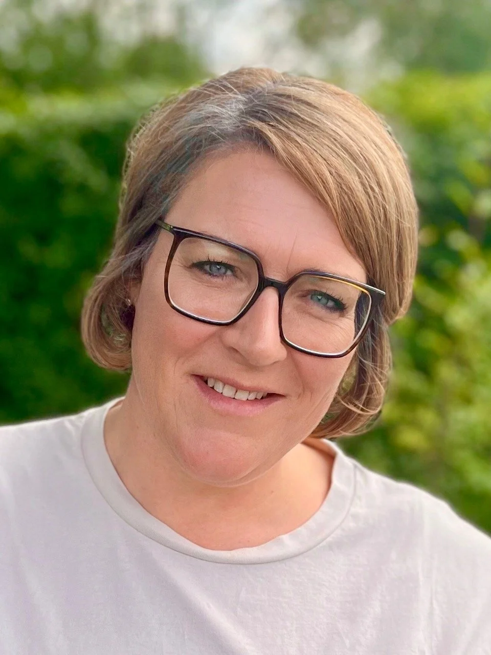 Nahaufnahme eines lachenden Frau mit Kurzhaarfrisur, Brille und weißem T-Shirt, im Grünen