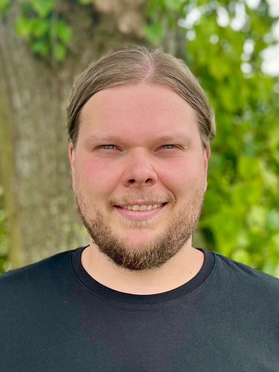 Ein Mann mit langem, glattem Haar und Bart steht vor einem Baum, umgeben von grünen Blättern, und lächelt in die Kamera.