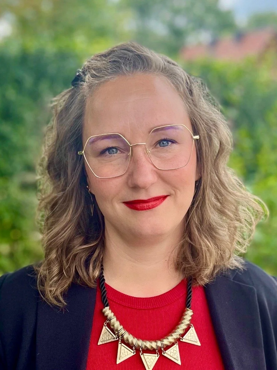 Nahaufnahme einer lächelnden Frau mit lockigem, braunem Haar, rotem Lippenstift, großen Brillengläsern, rotem Oberteil, schwarzer Jacke und einer auffälligen Ketten um den Hals, vor einer grünen, unscharfen Naturkulisse.