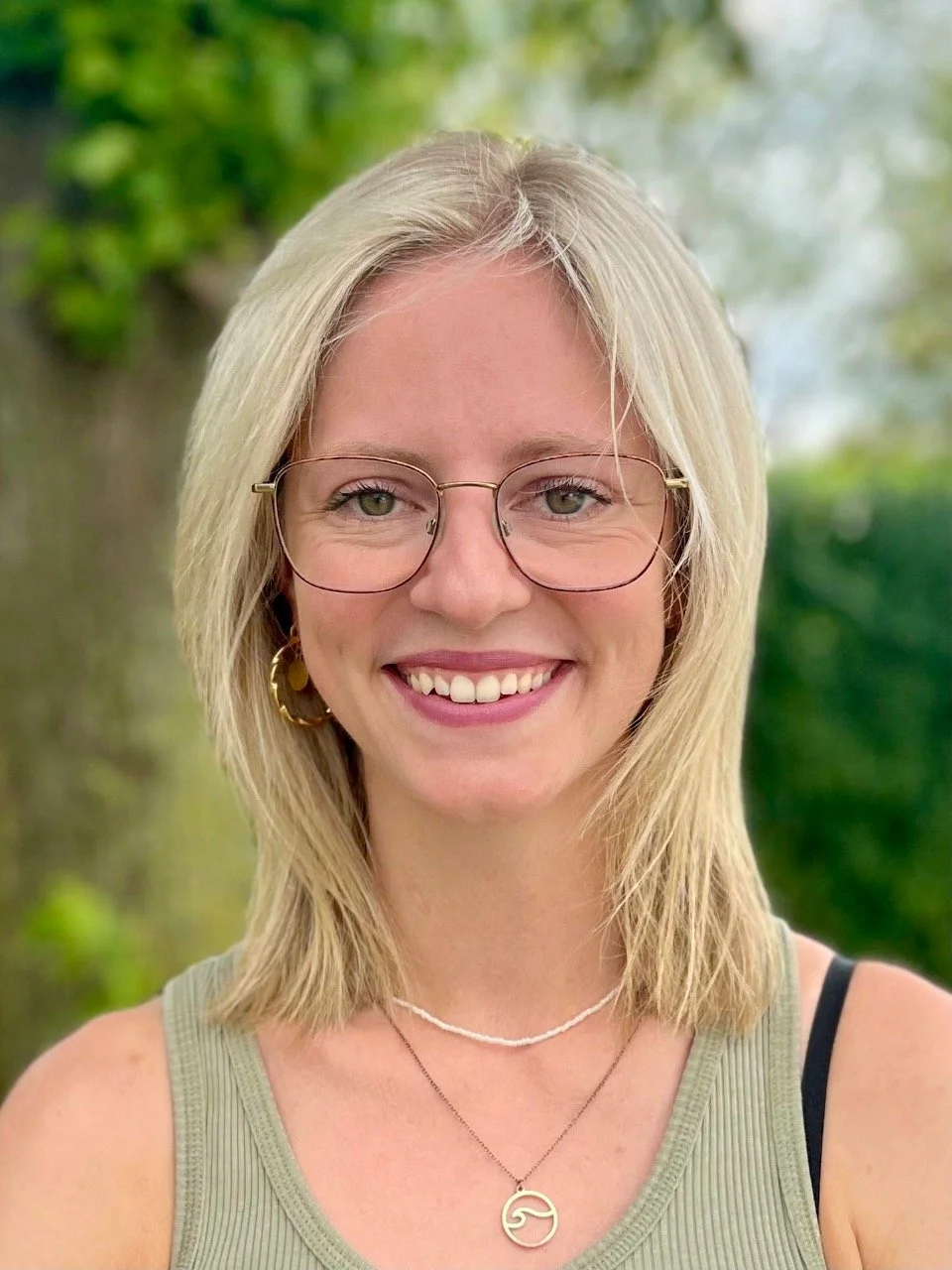 Nahaufnahme einer lachenden jungen Frau mit blondem, schulterlangem Haar, Brille, goldenen Ohrringen, Halsketten und lockeren Kleidung, vor einem grünen, verschwommenen Hintergrund.
