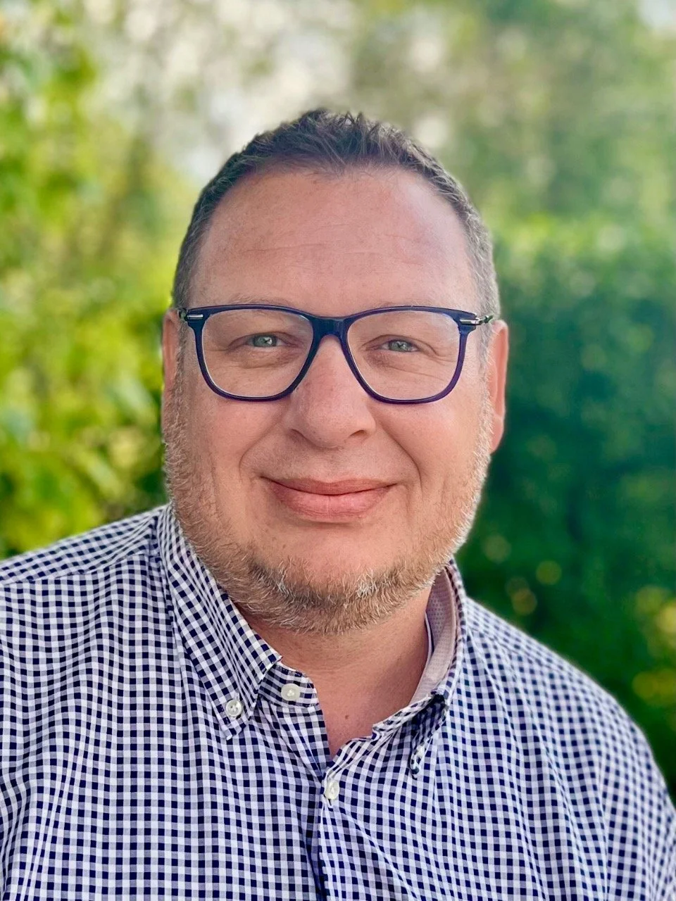 Ein Mann trägt eine Brille und ein kariertes Hemd, lächelt und steht vor einem grünen, bewaldeten Hintergrund.