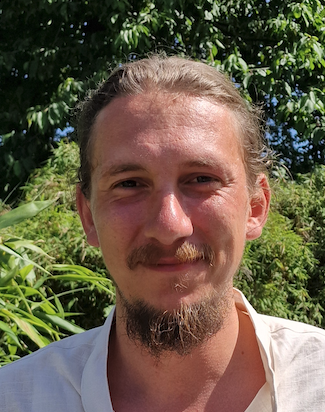 Ein Mann mit dunklen, nach hinten gegelten Haaren und blauen Augen, trägt ein schwarzes Polohemd, vor einem grünen, bewaldeten Hintergrund.