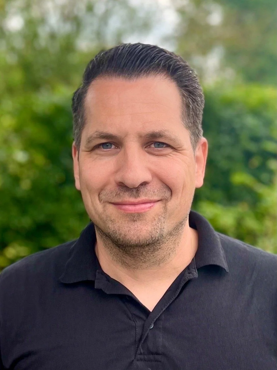 Ein Mann mit schwarzen Haaren und blauen Augen trägt ein schwarzes Polo-Shirt, steht vor grüner, bewaldeter Hintergrund, lächelt freundlich.