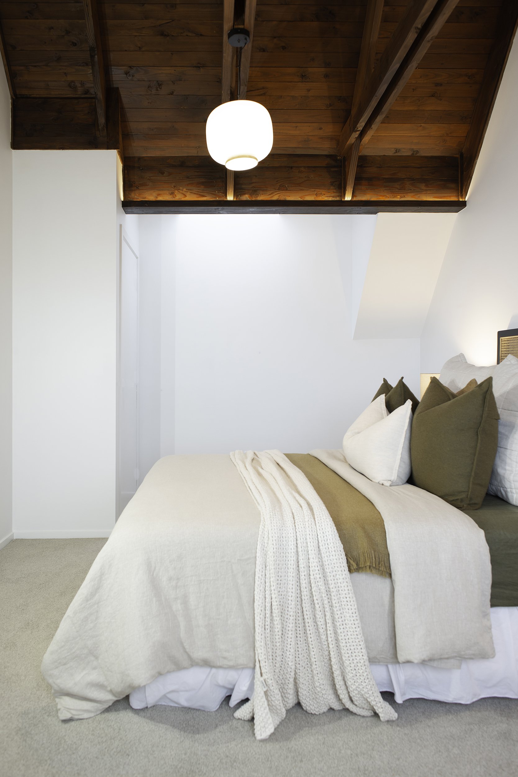 A cozy bedroom with a bed dressed in beige and white linens, multiple pillows, and a white knit throw blanket. The room has white walls, beige carpet, and a wooden ceiling with a hanging light fixture.