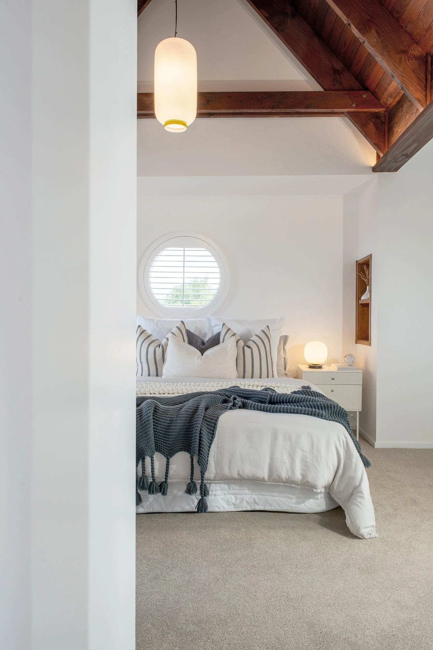A cozy bedroom with a white bed, decorated with striped and solid pillows, a dark blue throw blanket, and a small round window with blinds. A white nightstand with a glowing lamp and decorative items sits beside the bed, with wooden beams and ceiling above.