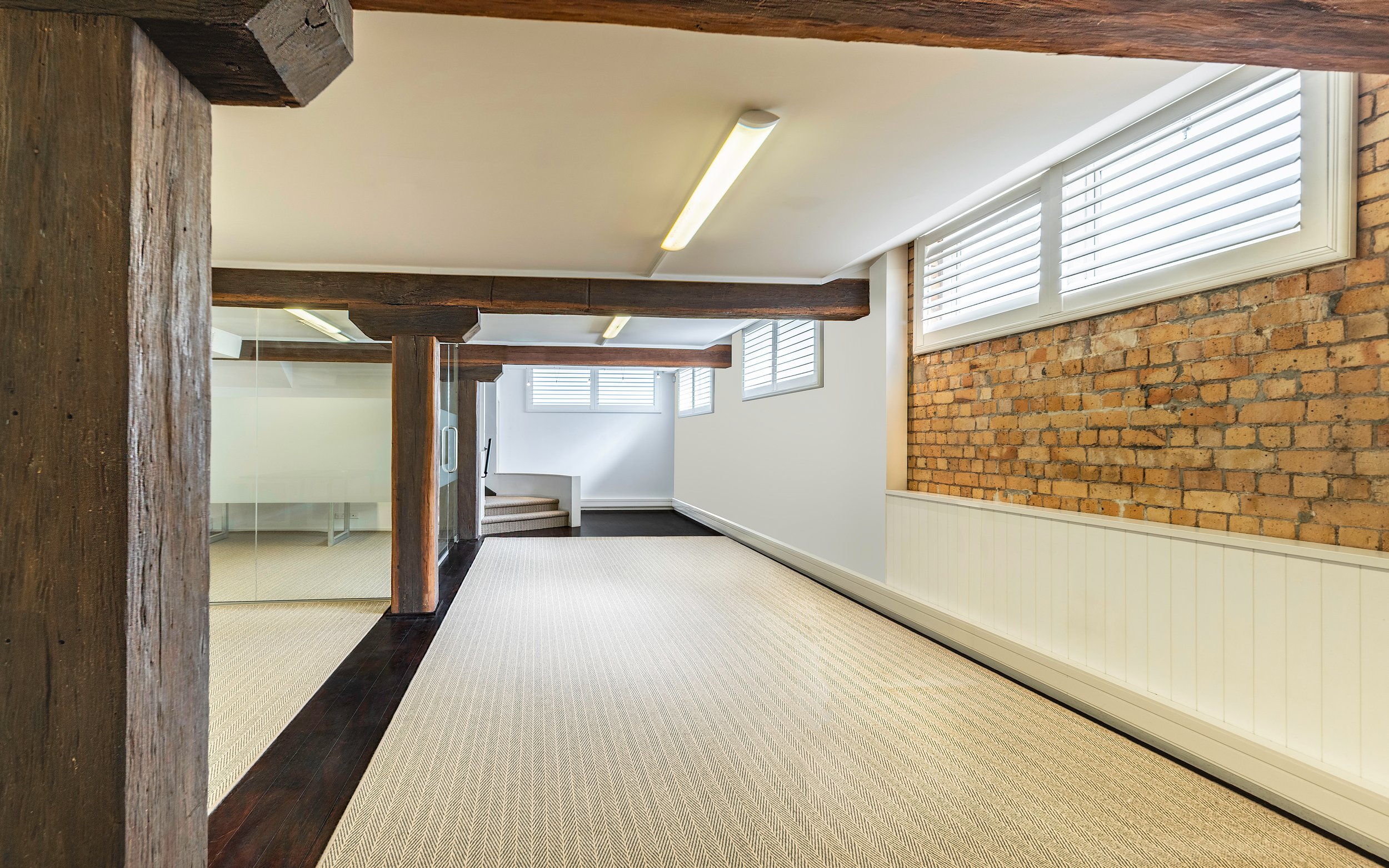Empty hallway with white walls, brick accent wall, wooden beams, carpeted floor, and windows with blinds letting in natural light.
