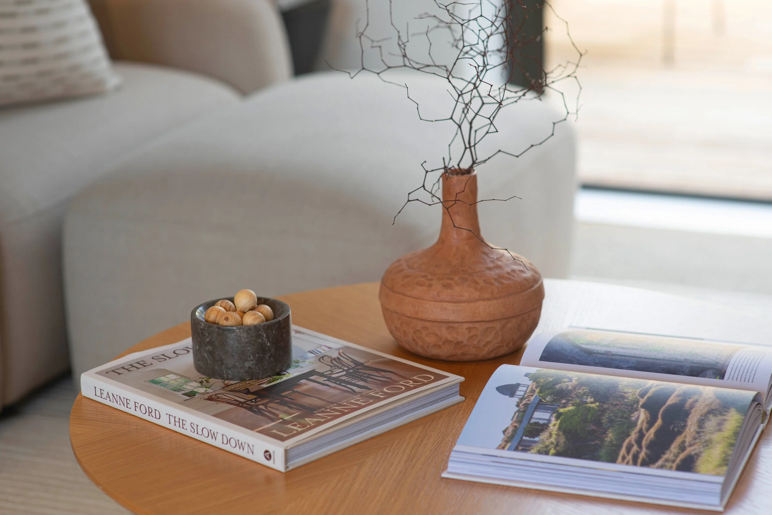 A round wooden table with a stack of books, a black bowl with wooden balls, and a large terracotta vase with dried twigs. The open magazine shows scenic landscape photographs. In the background, there is a beige sofa with a pillow.