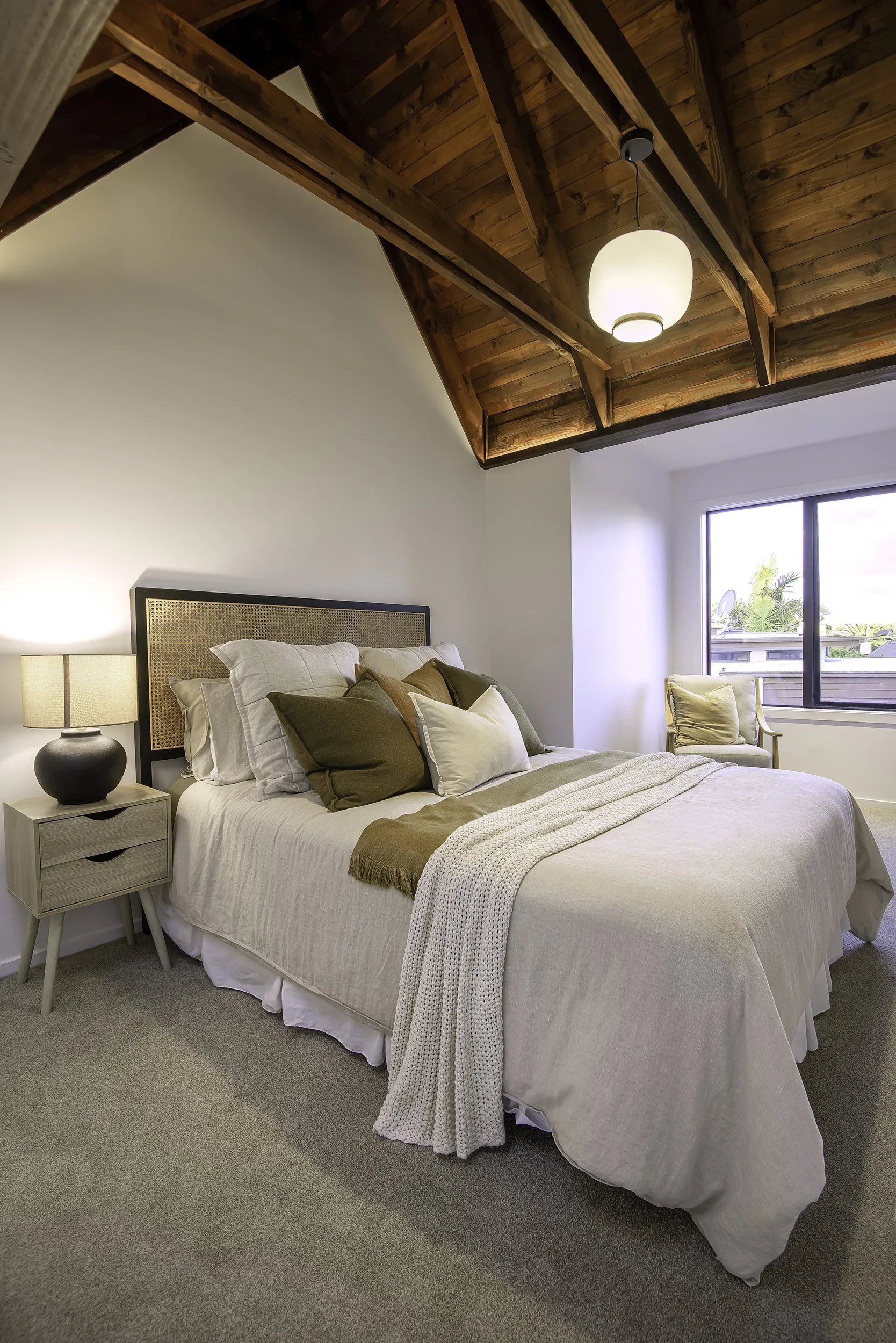 A cozy bedroom with a large bed covered in beige linen bedding, multiple pillows in earth tones, a wooden nightstand with a black lamp, an armchair by the window, and a wooden vaulted ceiling with a hanging light fixture.
