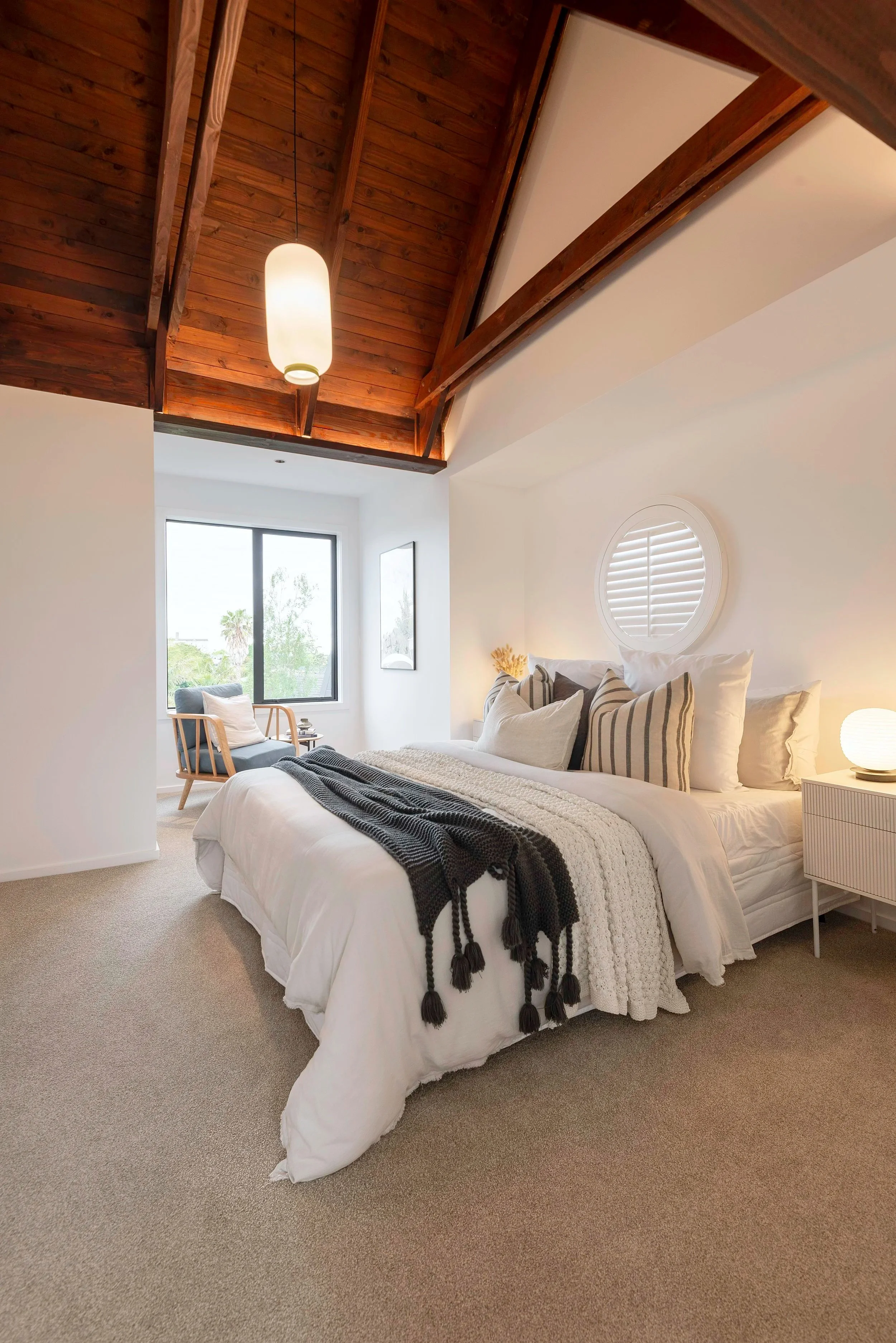 A cozy bedroom with a sloped wooden ceiling, white walls, and a large bed with white linens, multiple pillows, and a dark throw blanket. There is a circular window above the bed, a bedside table with a lamp, a picture on the wall, and a sitting area near a window with greenery outside.