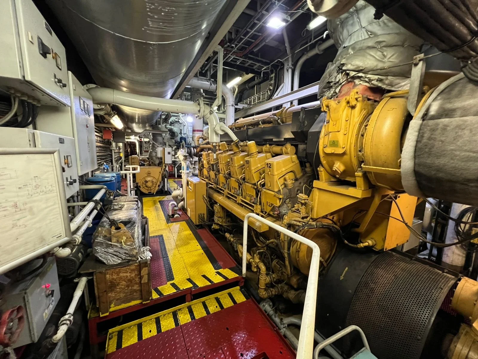 A large industrial engine room featuring a yellow Caterpillar generator, various pipes, control panels, and industrial equipment, with a walkway and safety rails.