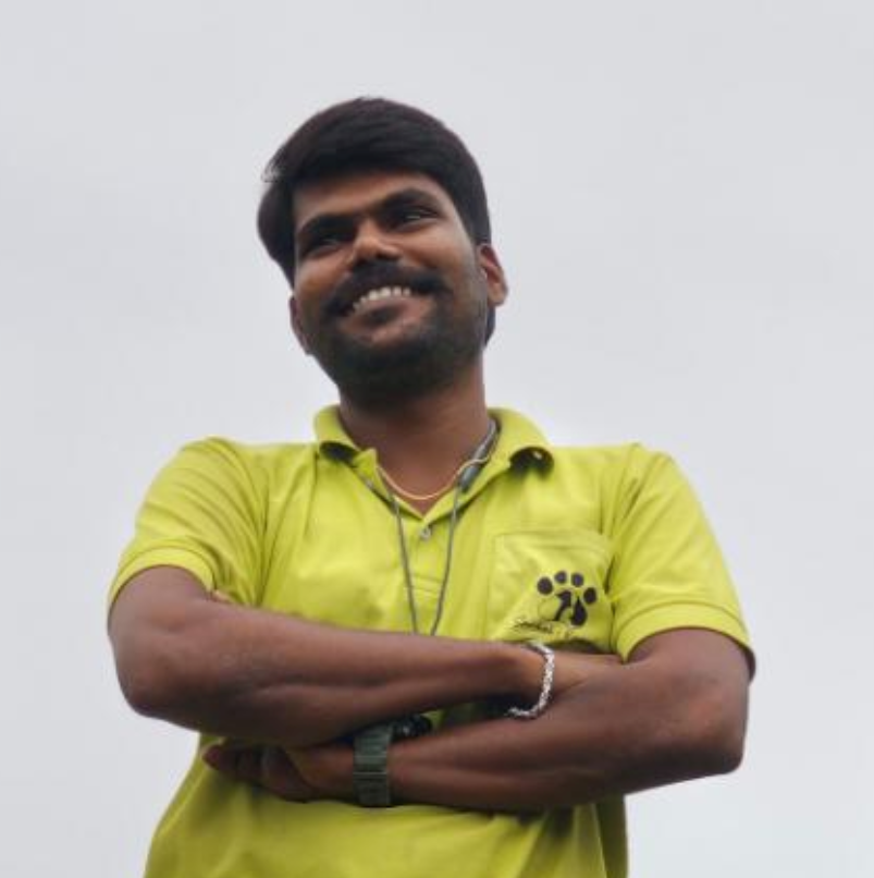 A smiling man with dark hair and beard wearing a yellow polo shirt with a paw print logo, standing outdoors against a light gray sky.