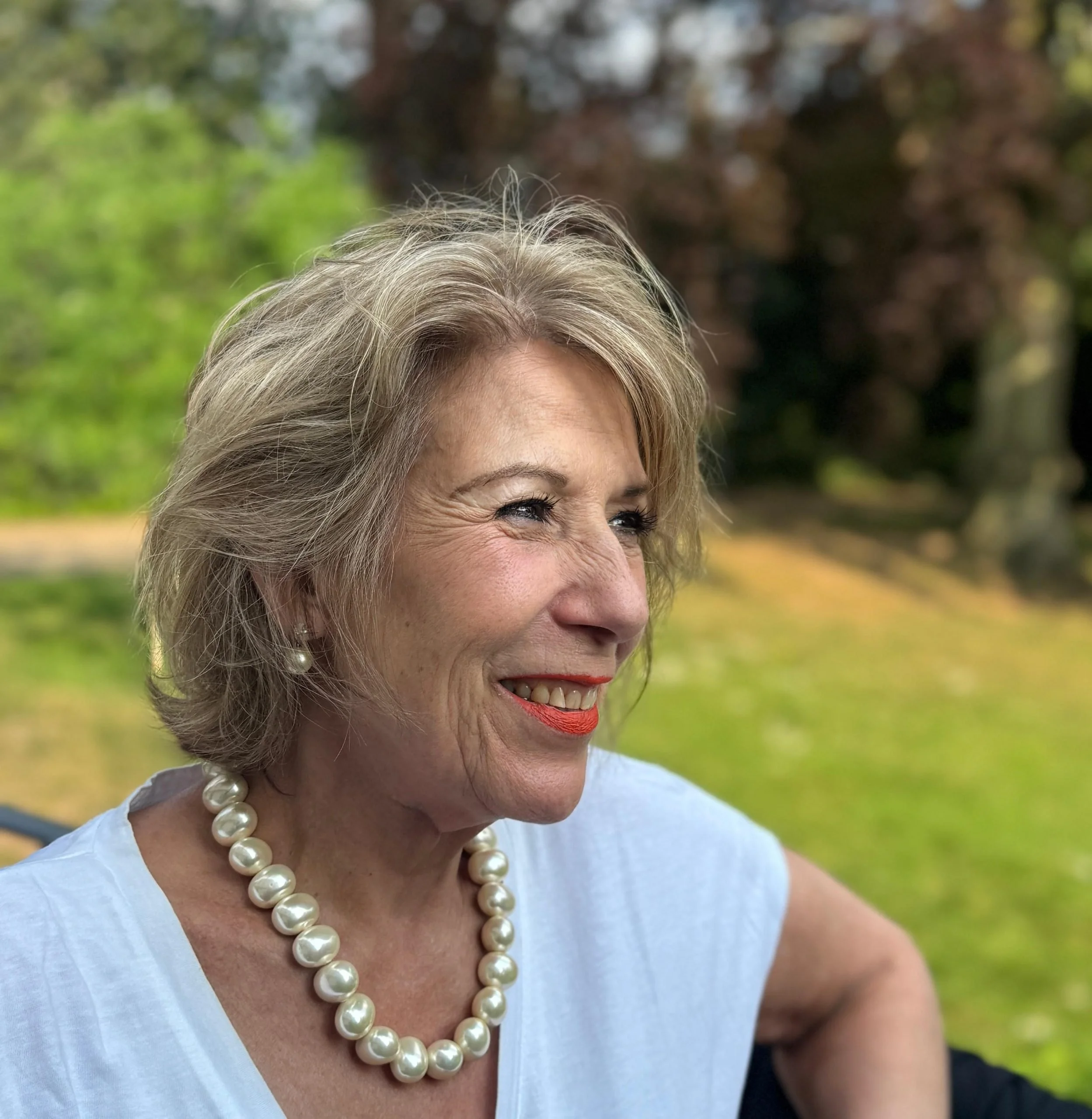 A woman with short blonde hair smiling, wearing a white top, pearl necklace, and pearl earrings, outdoors on a sunny day with trees and grass in the background.