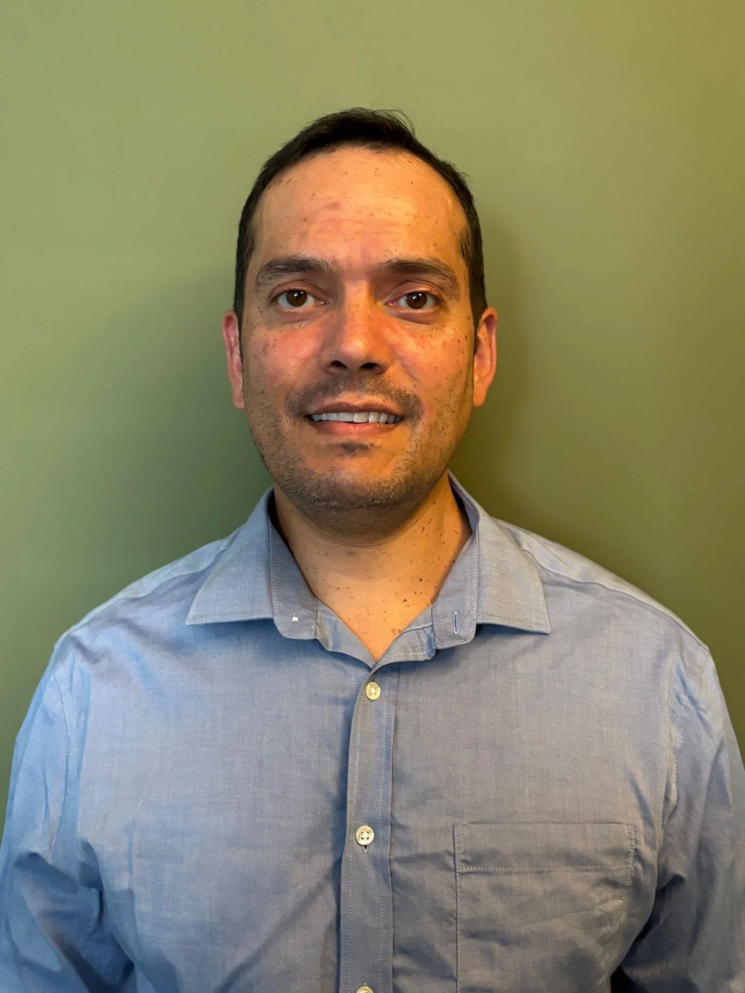 Portrait of a man with short dark hair, dressed in a light blue button-up shirt, standing against a plain green wall.