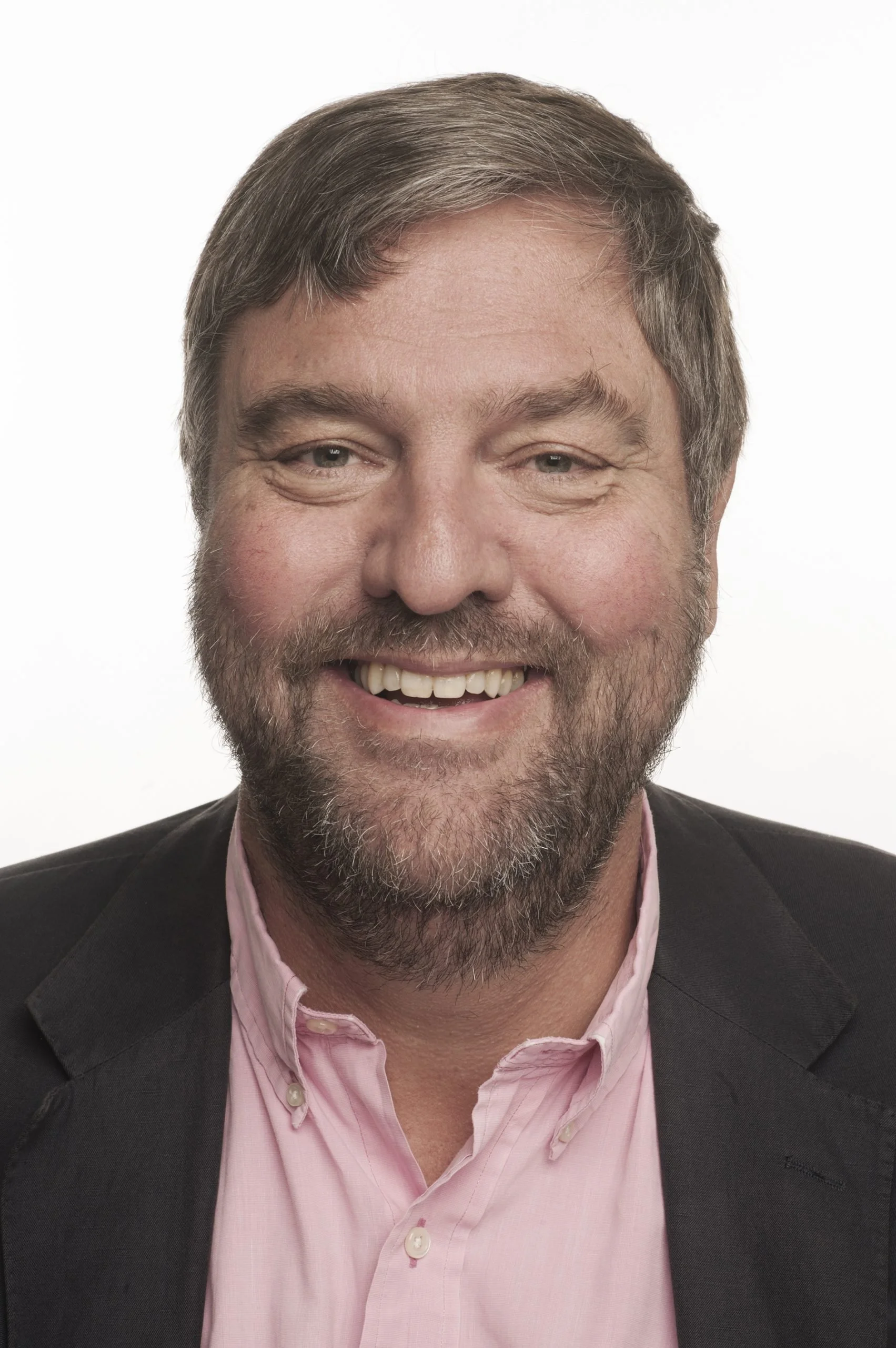 Headshot of a smiling man with gray hair, beard, wearing a black blazer and pink collared shirt, against a plain white background.