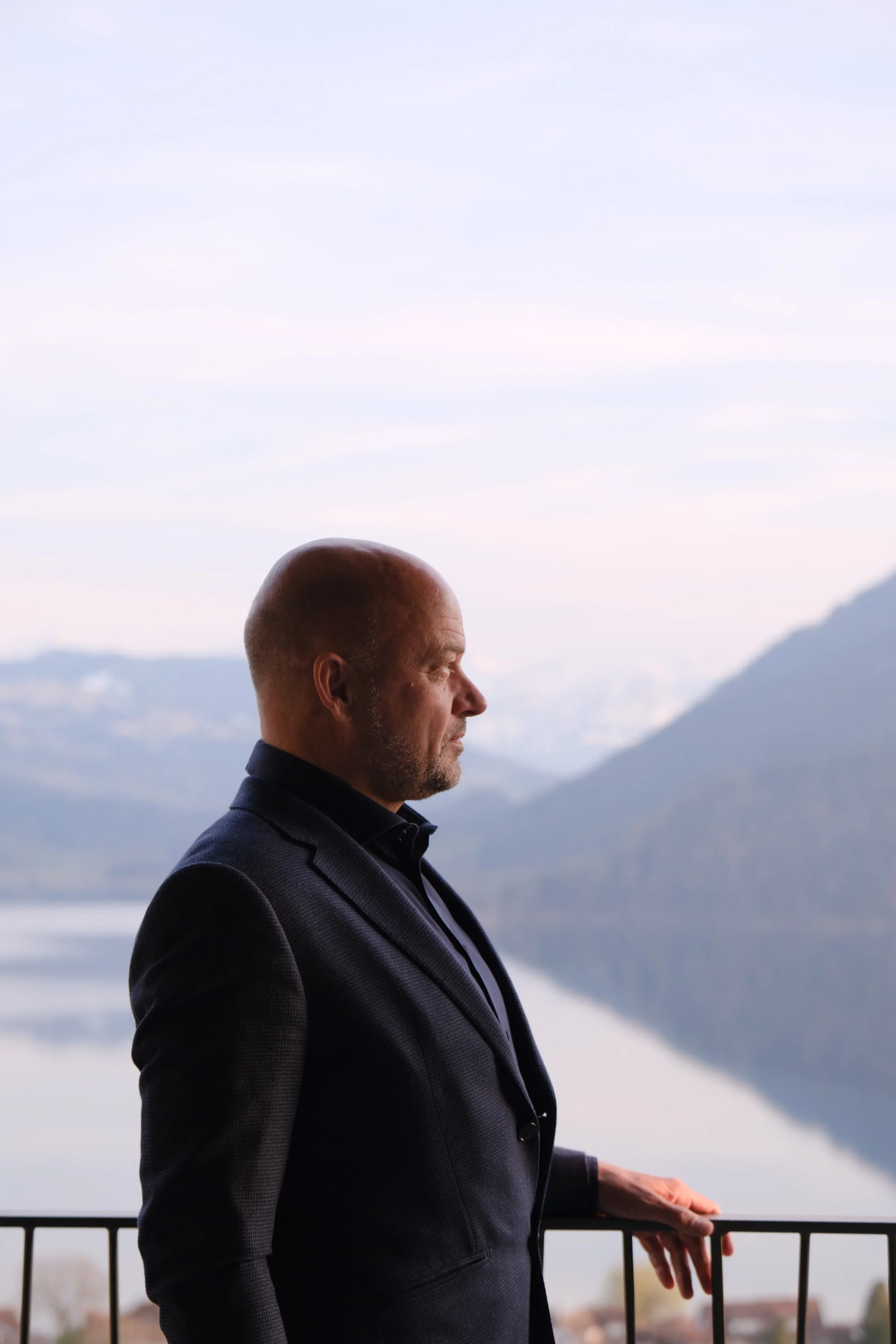 A man in a dark suit standing on a balcony overlooking a lake with mountains in the background.