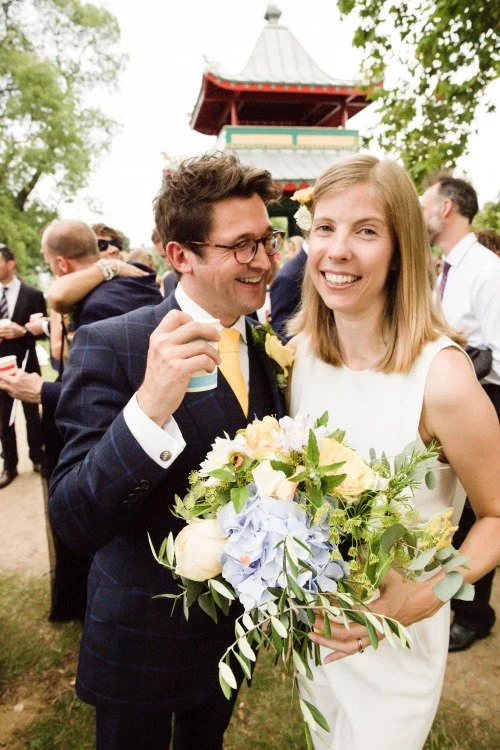 A smiling woman in a white dress holding a large bouquet of flowers at an outdoor wedding celebration, with a man in a dark suit and yellow tie standing beside her, both surrounded by other guests, trees, and a pagoda-style structure in the backgroun