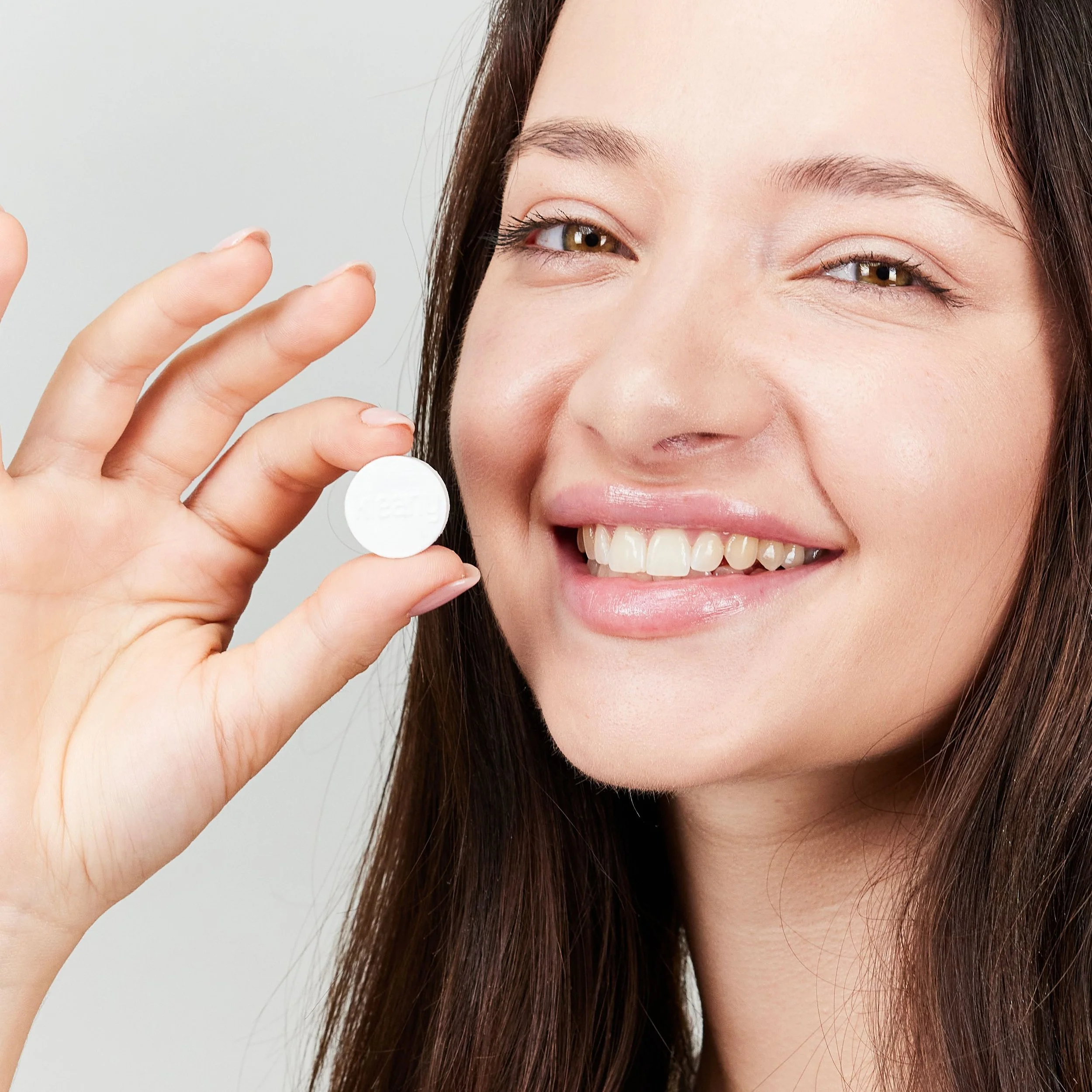 A woman smiling and holding a white pill between her thumb and index finger.