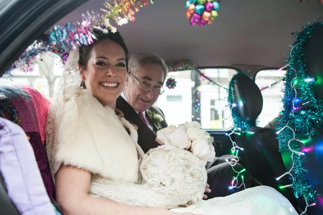 A bride and an older man, possibly her father, sitting inside a decorated vehicle, smiling. The bride wears a white fur stole, lace dress, and veil, holding a bouquet of white flowers. The vehicle is decorated with Christmas tinsel and lights, with c