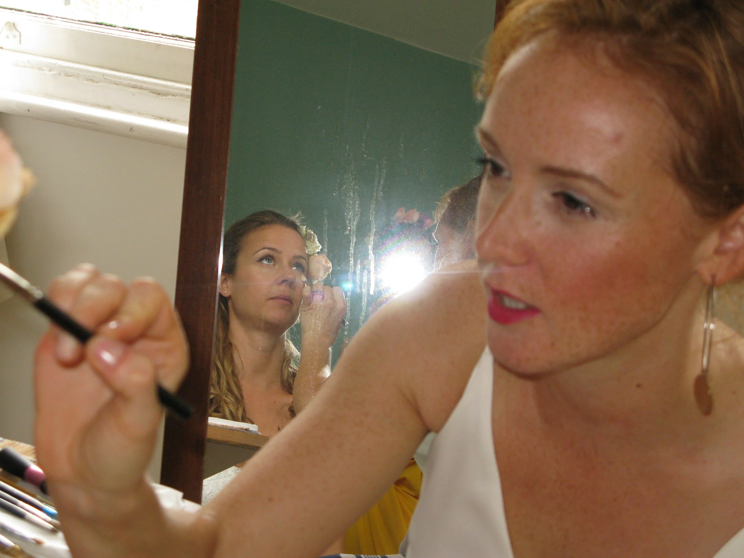 A woman with red hair and earrings applies makeup while looking into a mirror, with her reflection visible in the mirror behind her.