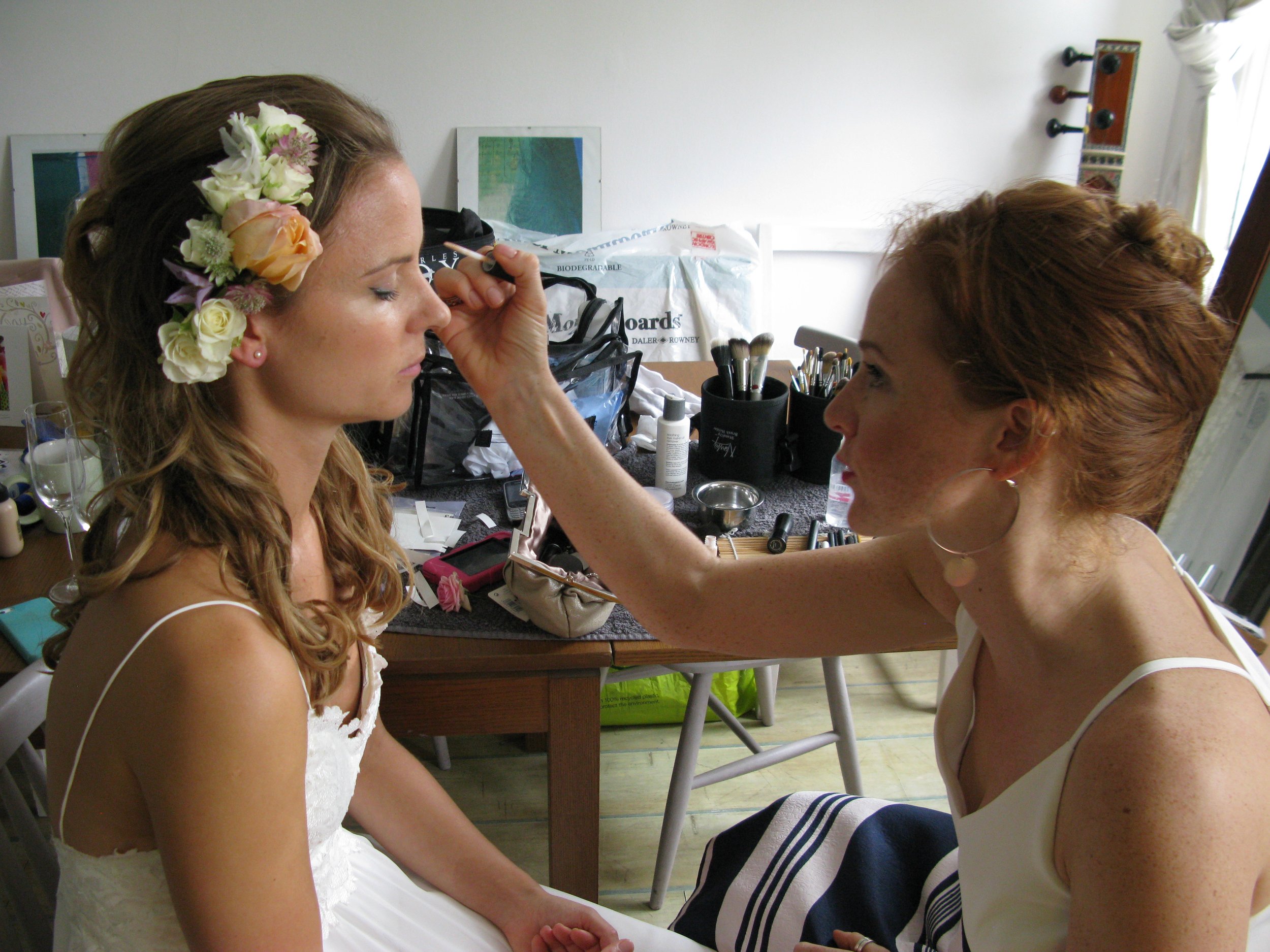 A woman with a flower headpiece gets her makeup done by another woman with large hoop earrings in a room with makeup tools and supplies on the table.
