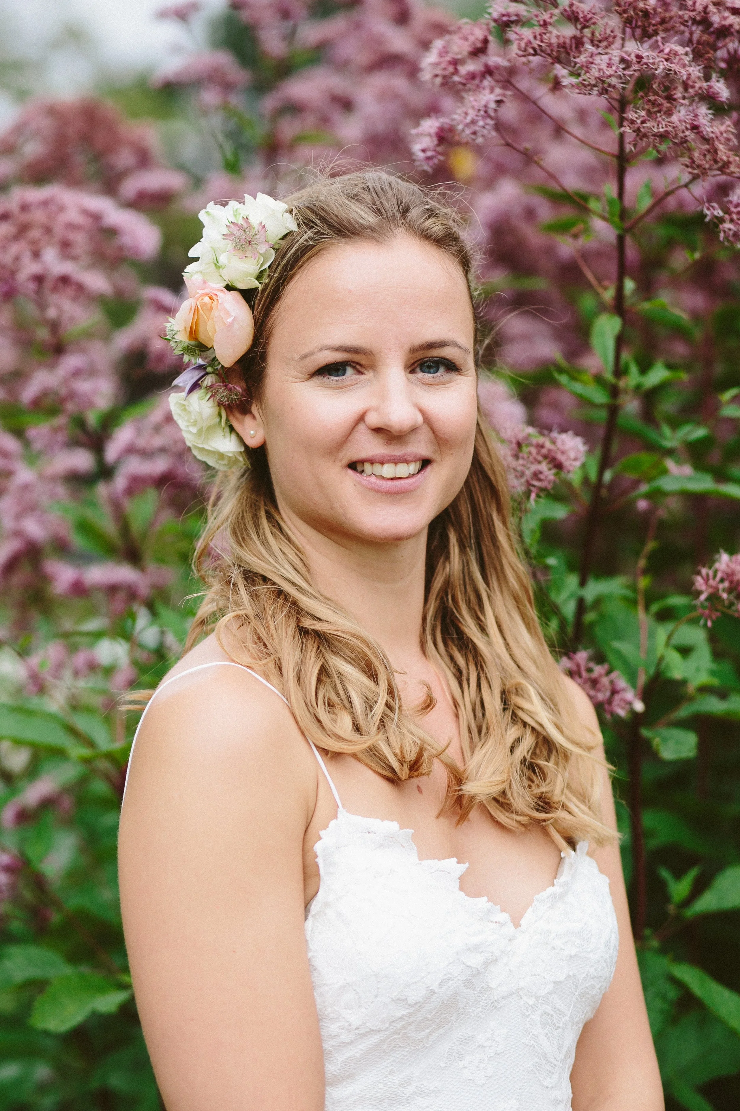 A woman with long, wavy hair and a floral headpiece, wearing a white lace dress, standing among pink flowering plants.