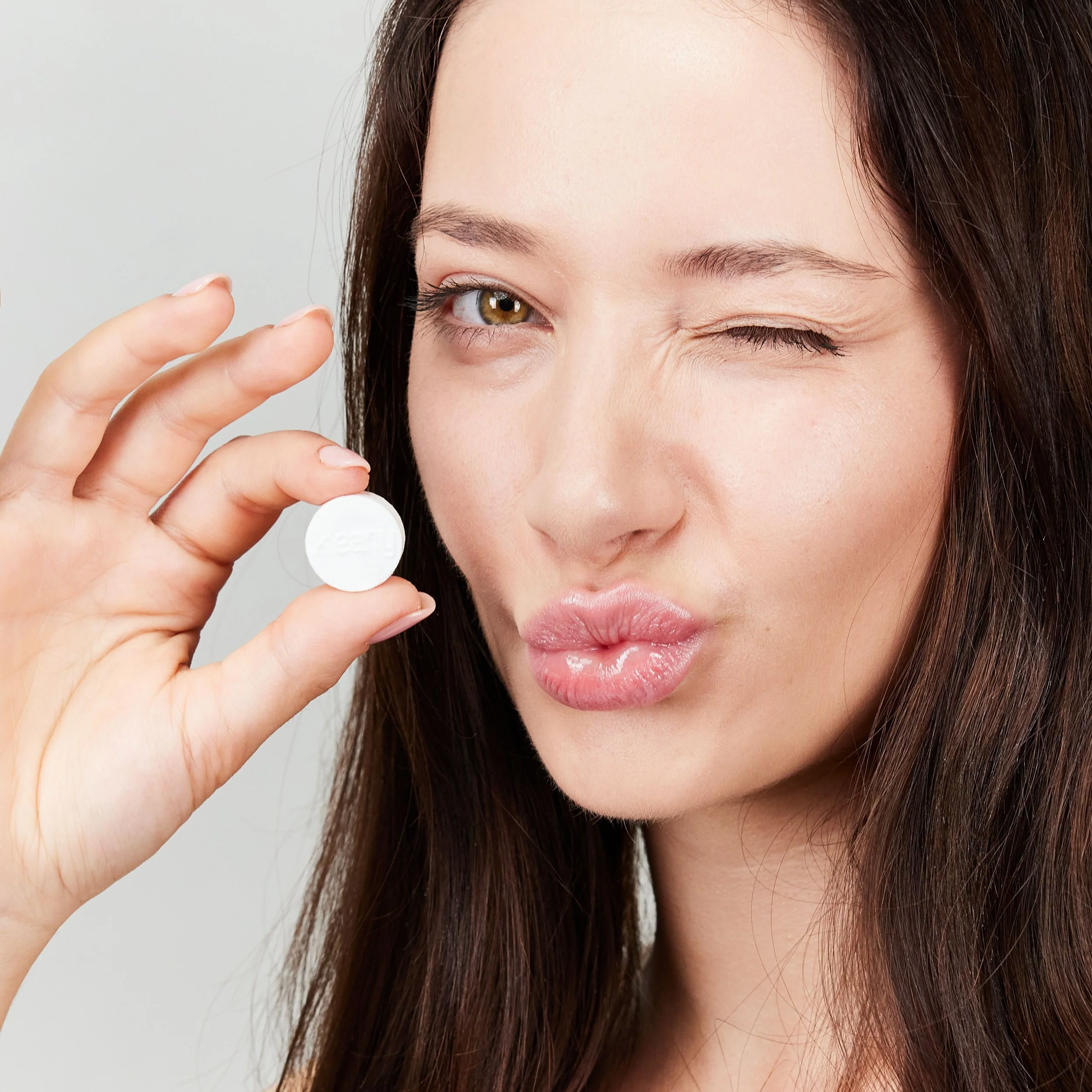 A woman with long brunette hair winks and holds a white pill close to her face.