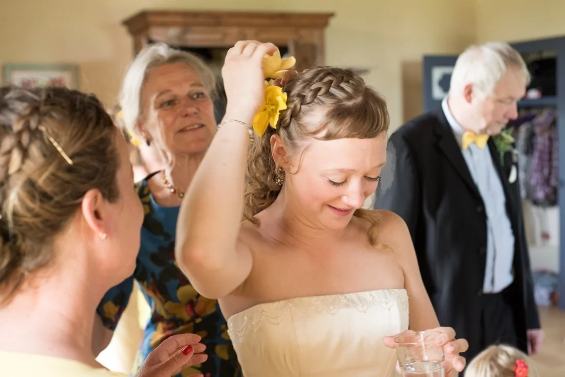 A young woman dressed in a strapless gown is smiling and holding a plastic cup, while another woman is placing a yellow flower in her hair. Two elderly people are in the background, one man and one woman, dressed formally.