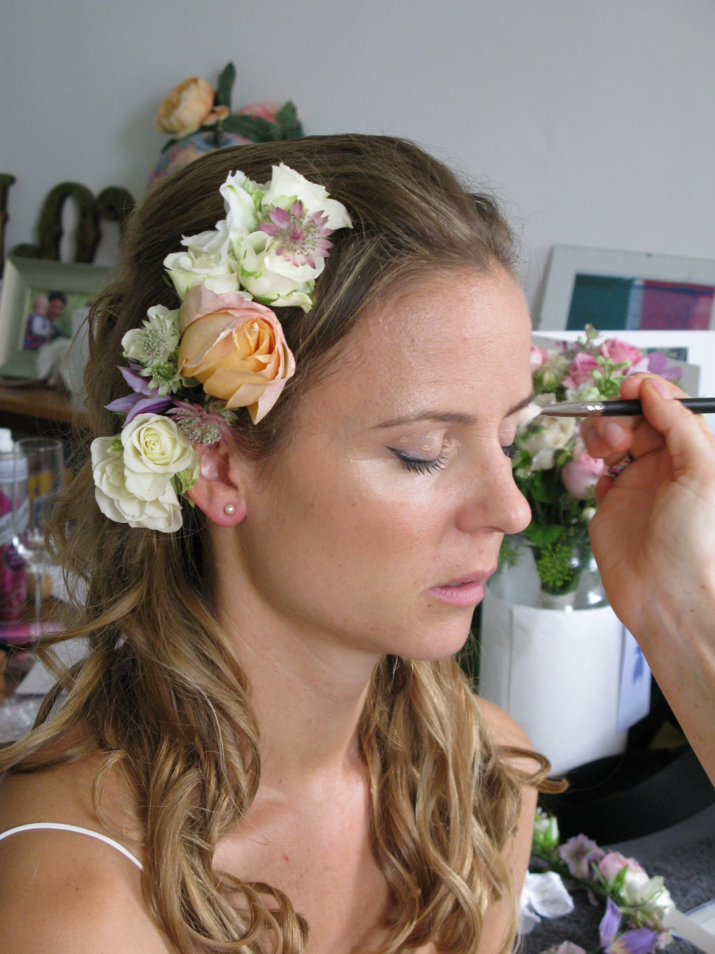 A woman with long blonde hair and floral headpiece having makeup applied by a makeup artist.