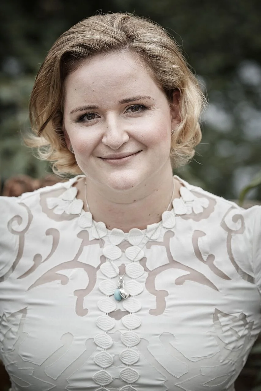 Close-up of a smiling woman with short, wavy blonde hair, wearing a white embroidered top and a layered necklace with circular and heart-shaped pendants, outdoors with blurred greenery in the background.
