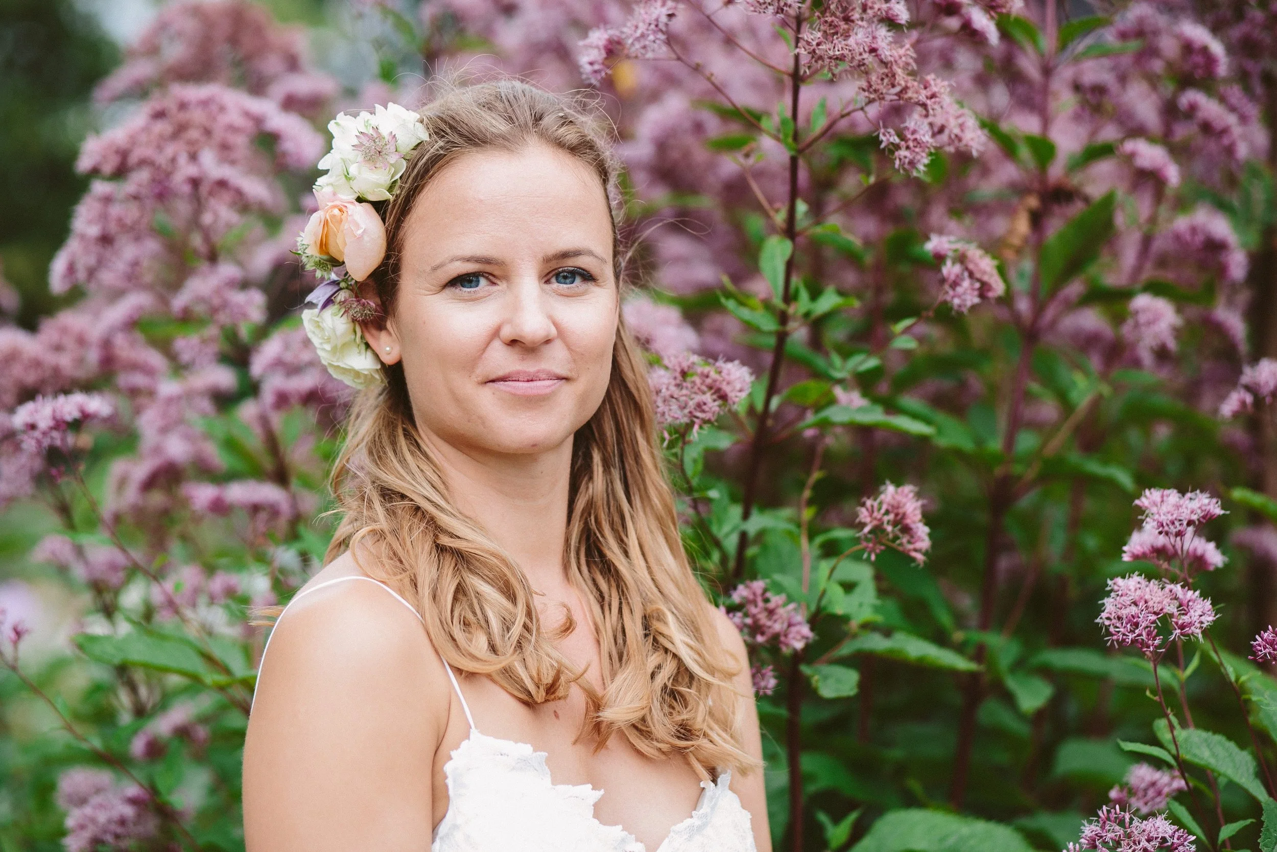 A woman with long, wavy blonde hair wearing a white dress and a floral crown, standing in front of pink blossoming flowers.