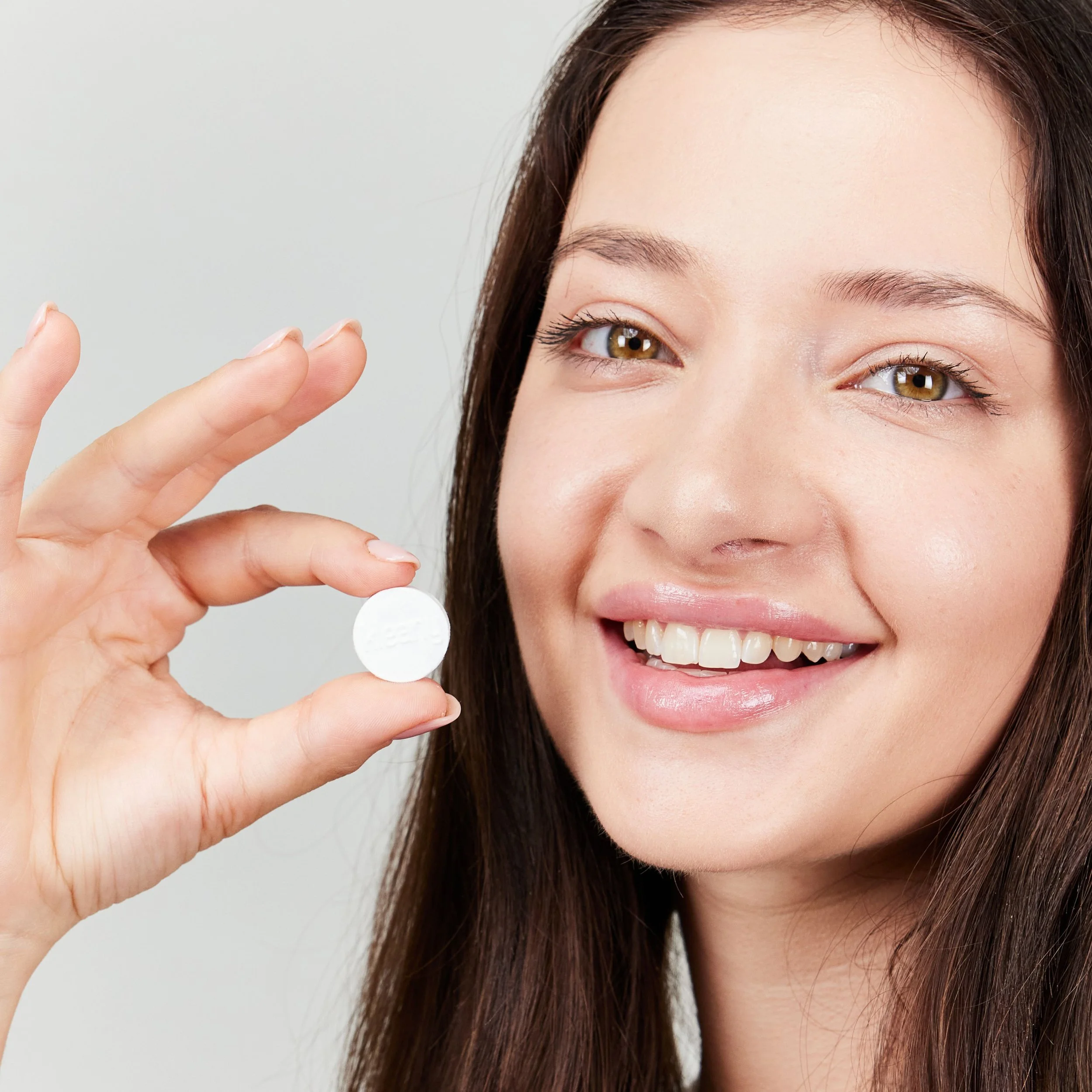 A smiling woman with brown hair holding a white pill close to her face.