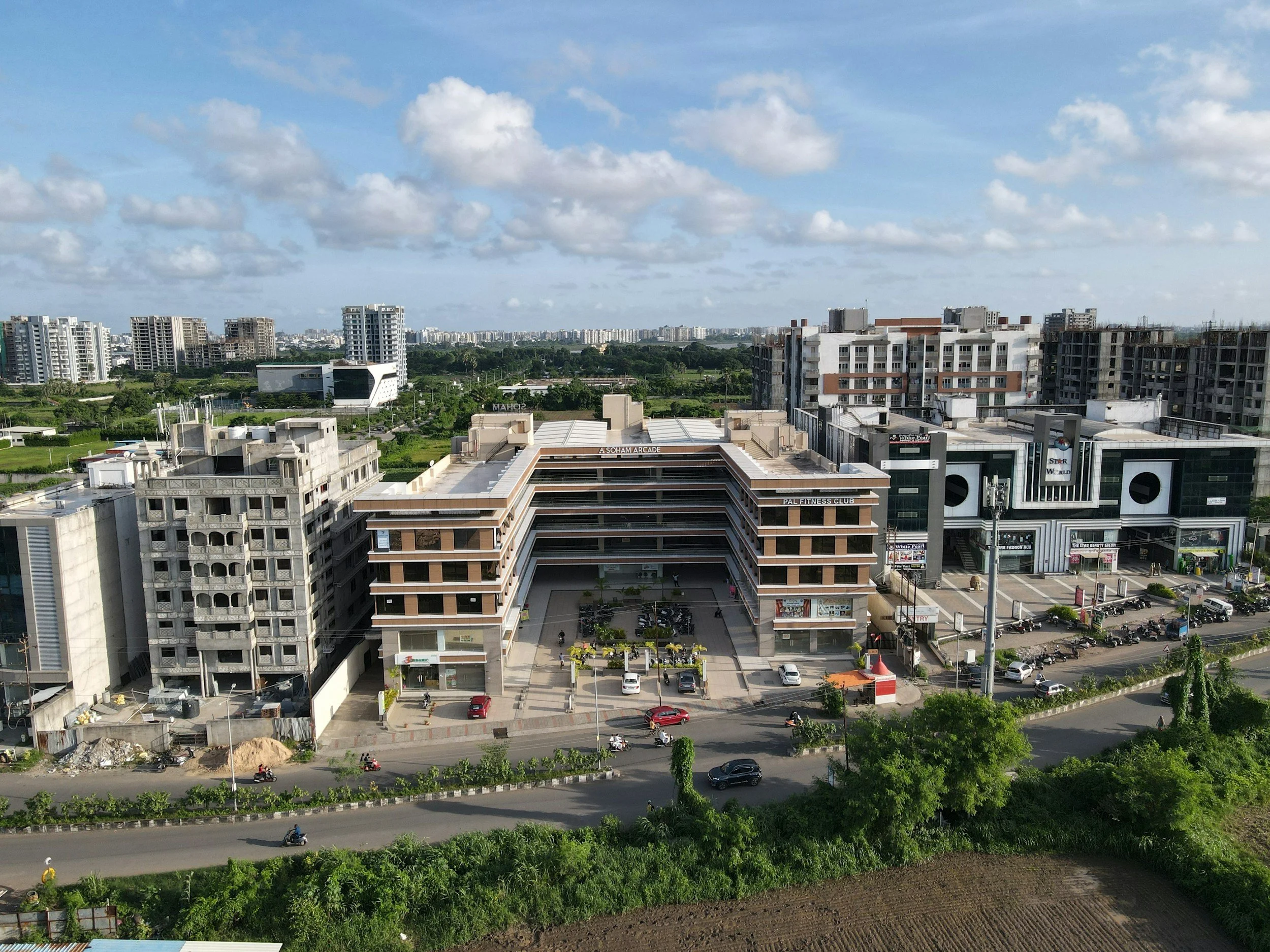 A cityscape featuring low-rise and mid-rise buildings, a shopping mall with commercial establishments, surrounded by greenery and roads with moving vehicles, under a partly cloudy sky.