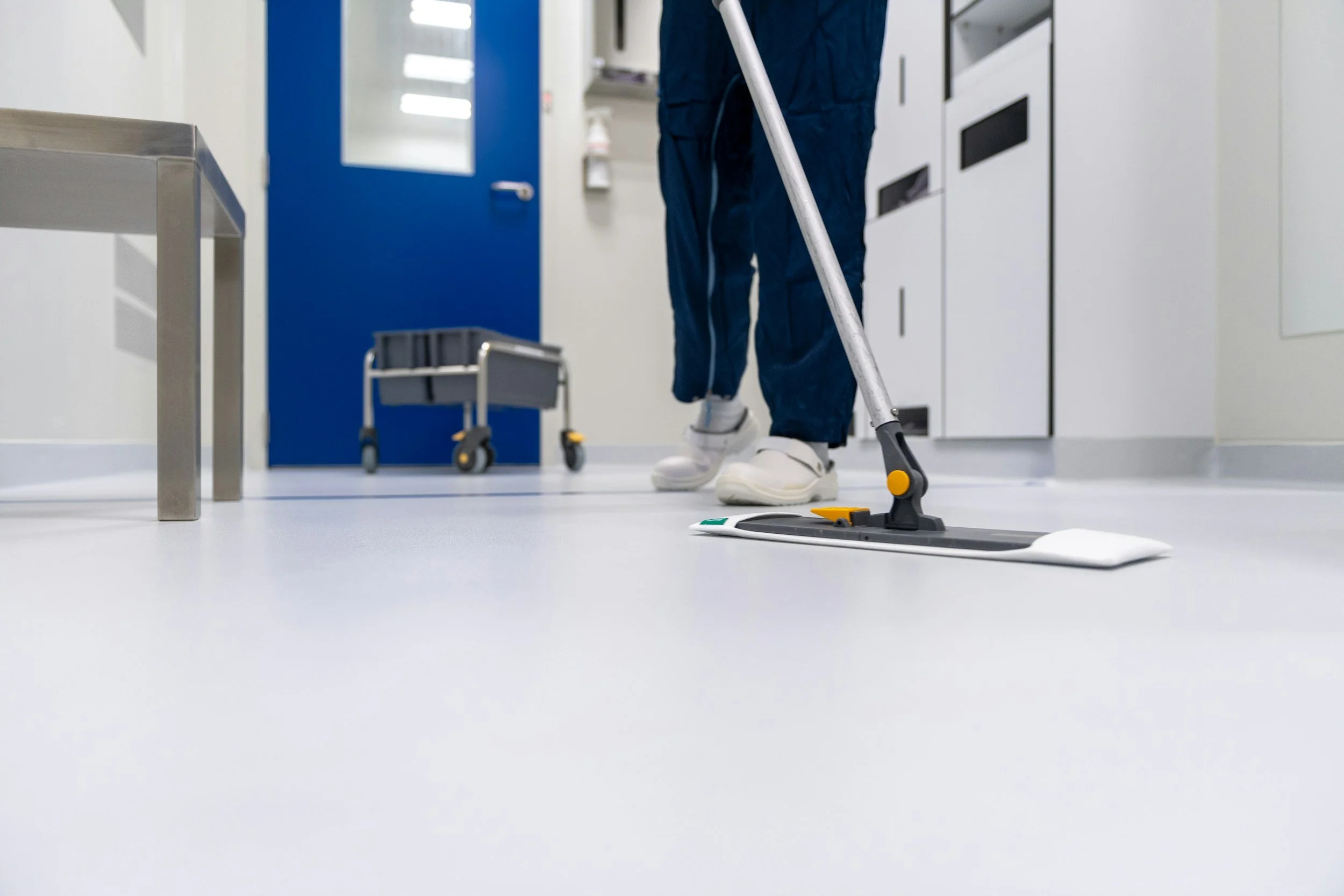 Person cleaning the floor with a mop in a sterile hospital or medical facility, white floors, blue door in the background, medical lockers, and equipment visible.