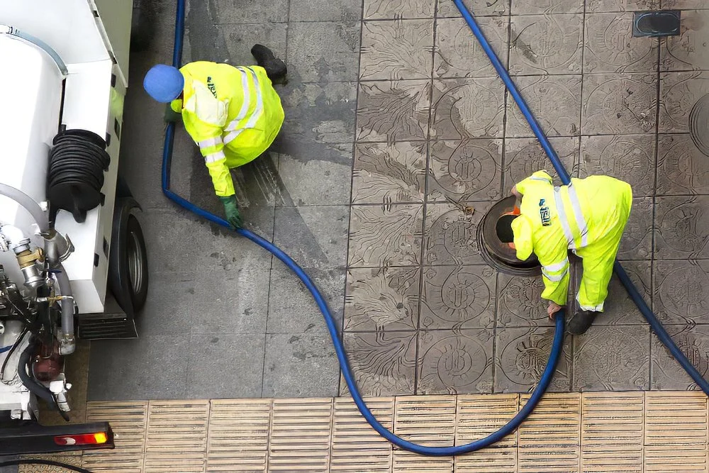 Two workers in yellow safety suits and blue helmets using hoses to clean a tiled sidewalk near a street.