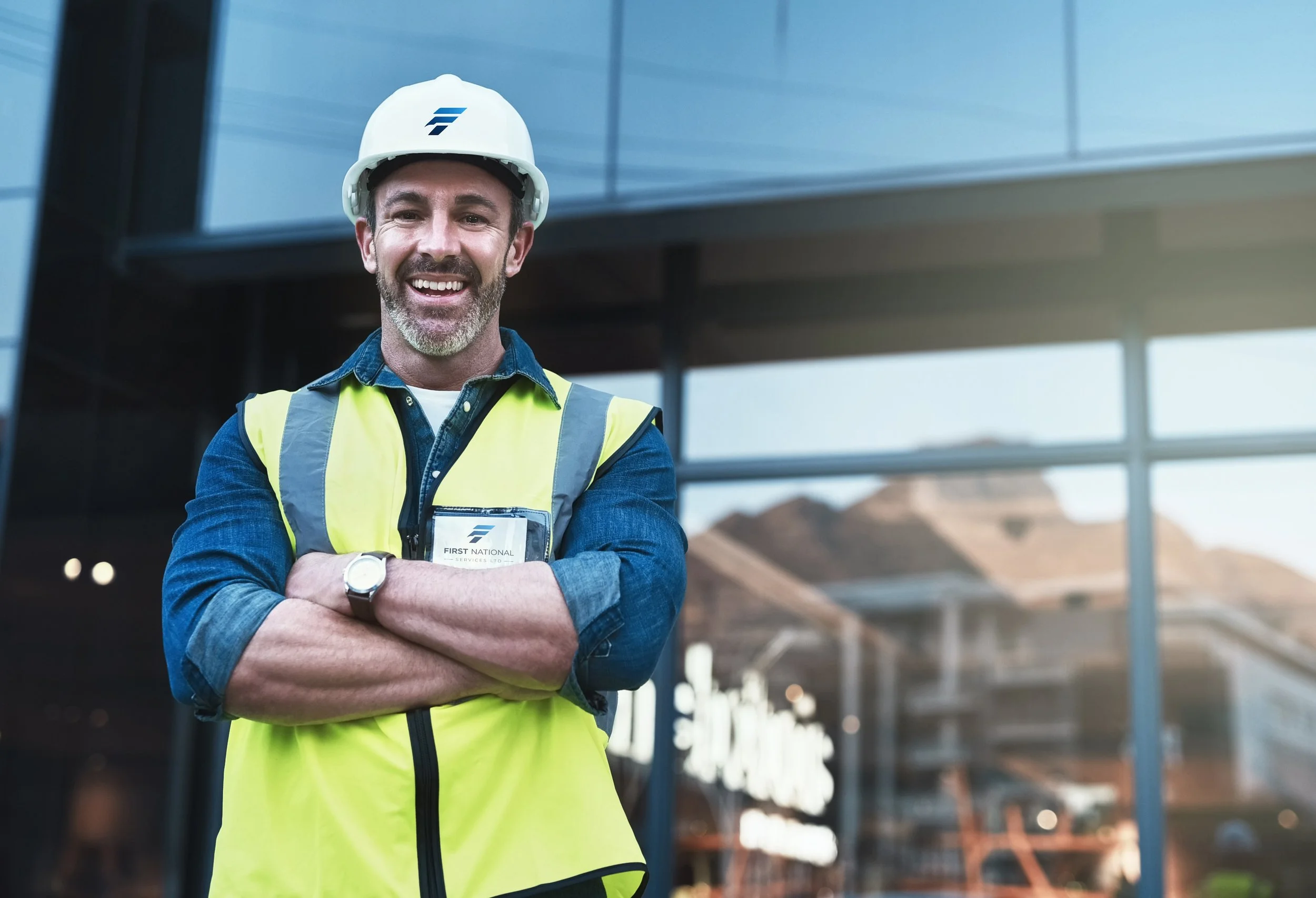 First National Services - A smiling construction worker with a beard, wearing a white safety helmet and yellow safety vest, standing with arms crossed outside a modern building with mountains in the background.
