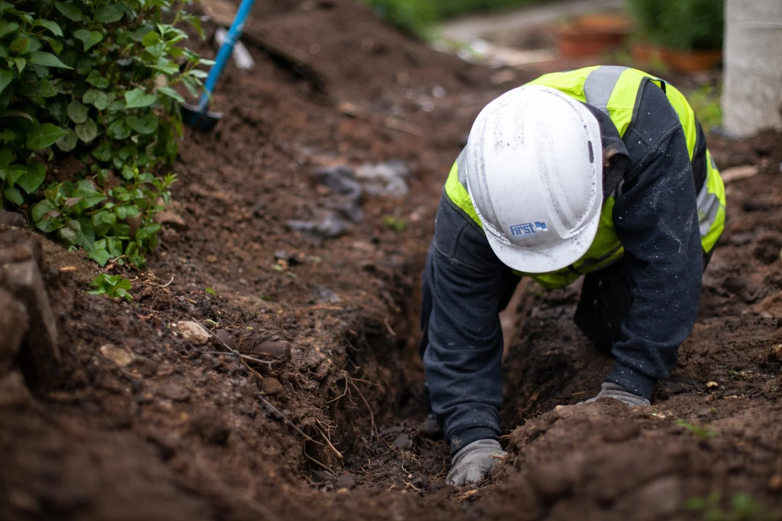 A construction worker wearing a white hard hat, a yellow safety vest, and dark clothing is planting or working in a garden bed with soil, surrounded by green plants.