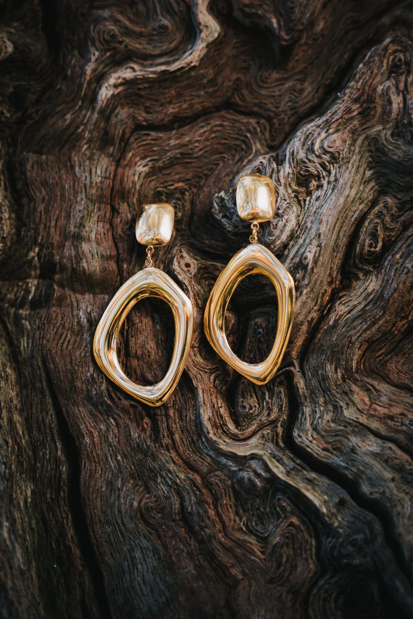 A pair of gold-colored hoop earrings with teardrop-shaped rings hanging from a dark, textured, wood background.