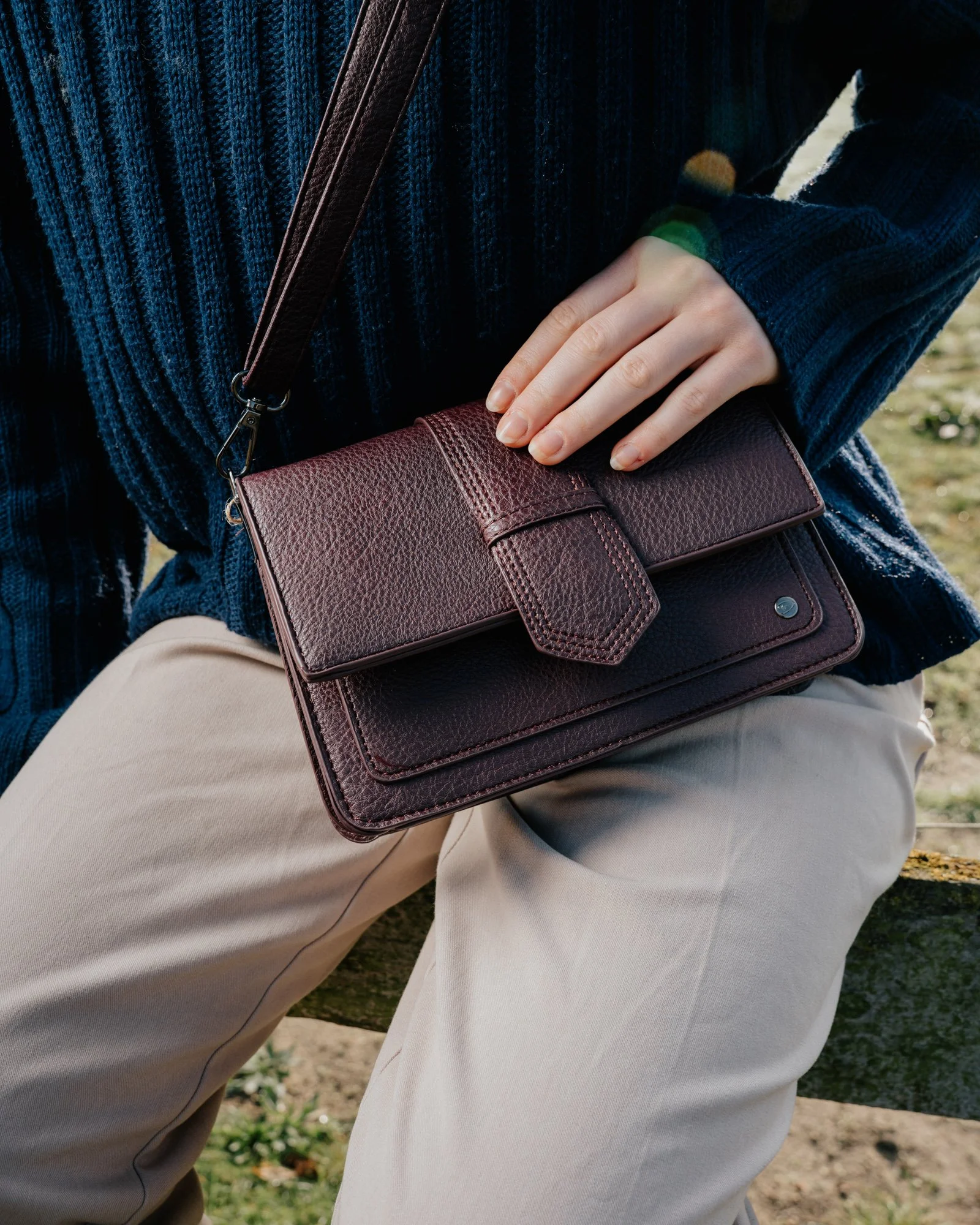 A person sitting outdoors, wearing a blue sweater and beige pants, holding a dark red leather purse with a strap.