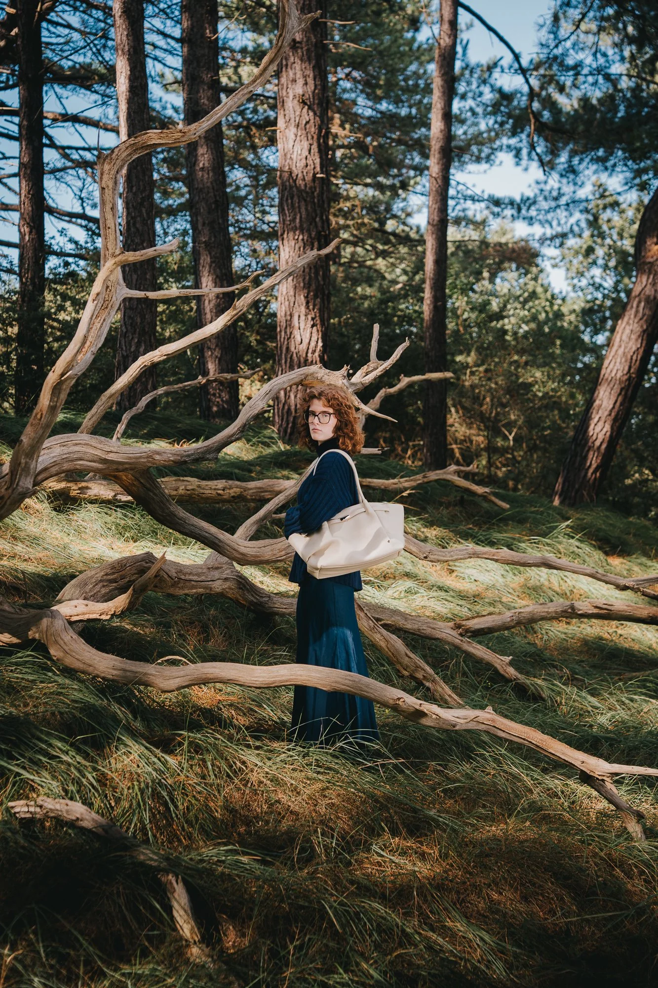 A woman with curly hair and glasses standing among fallen tree branches in a forest, wearing a dark blue outfit and carrying a beige bag.
